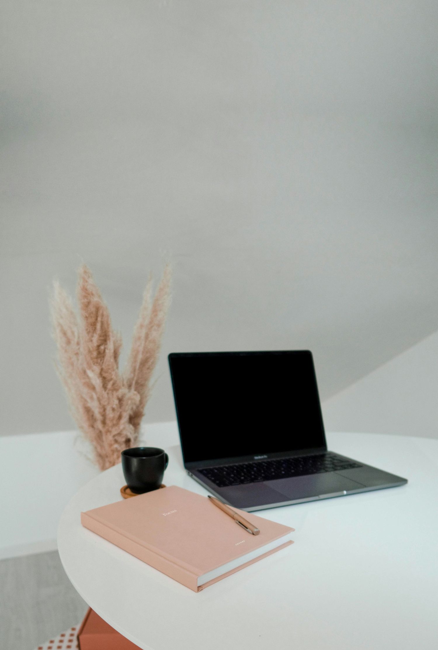 A minimalist desk setup with a laptop, pink notebook, and pampas grass decoration against a light gray wall.