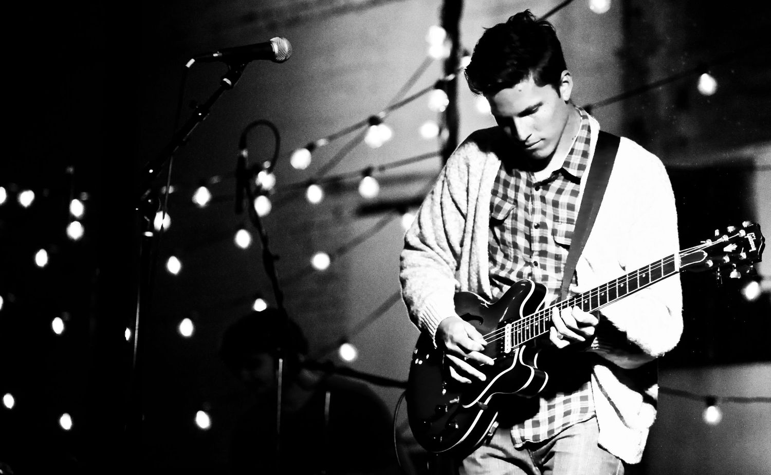 Black and white photo of a male musician playing an electric guitar on stage under spotlights, photographer Alec Watson