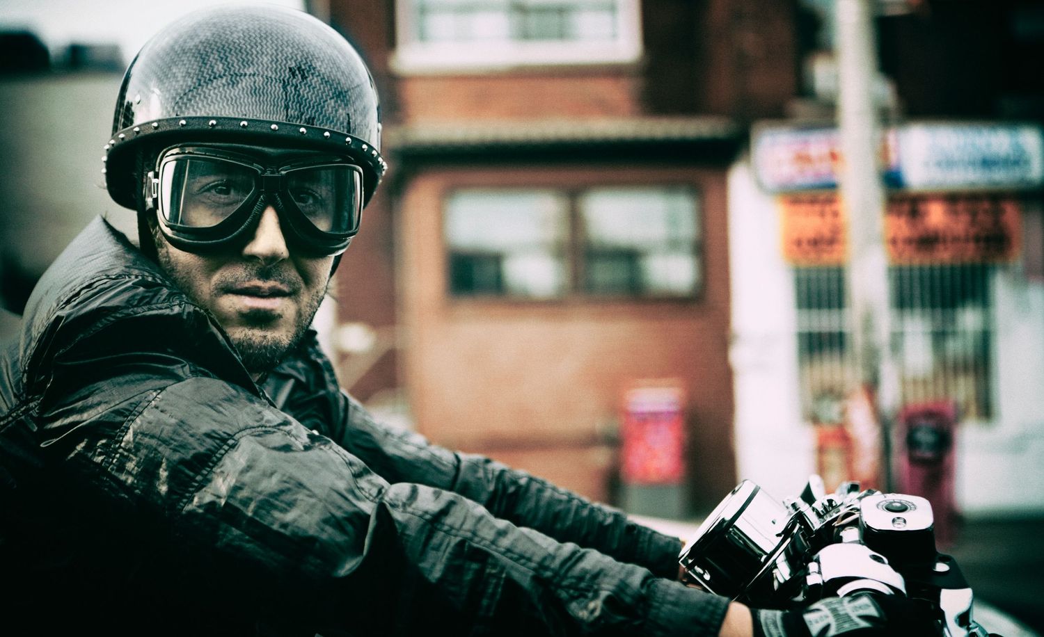 Portrait of a man riding a motorcycle wearing vintage goggles and helmet on a city street, Michael Crispel, photographer Alec Watson