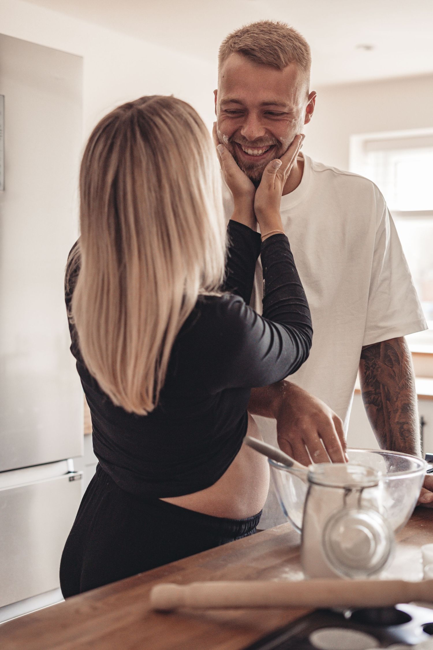 A couple shares an intimate moment in a bright kitchen while cooking together.
