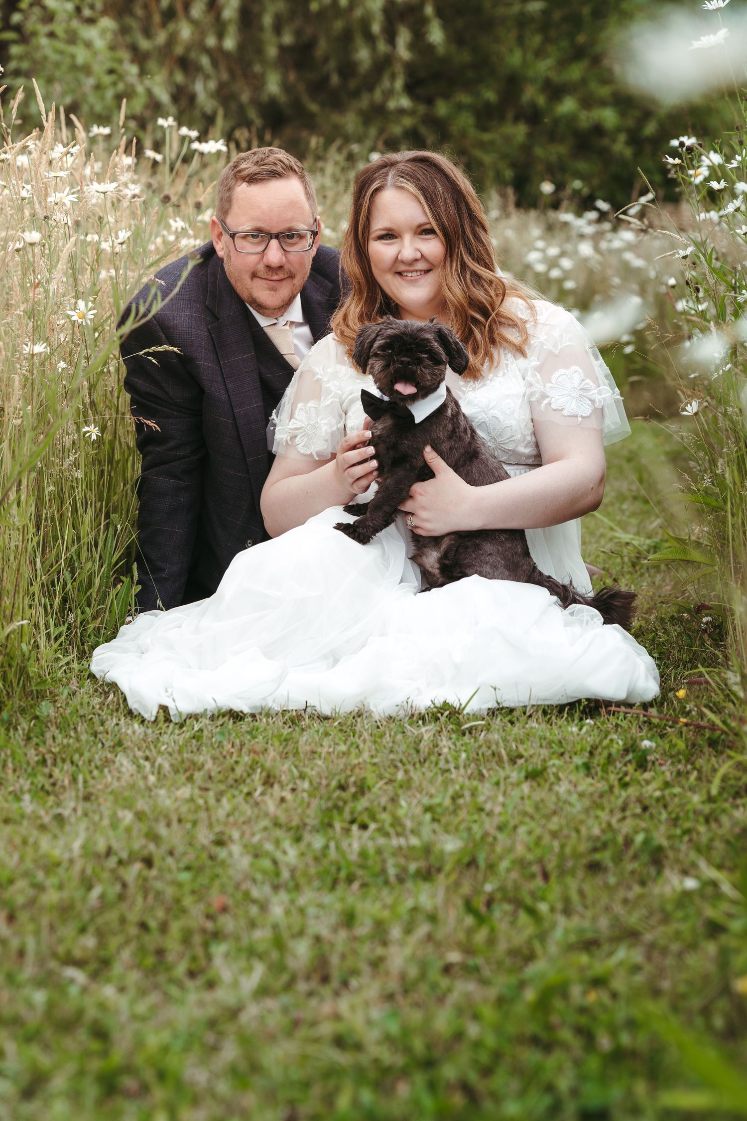 Wedding couple sits in wildflower meadow with their black dog.