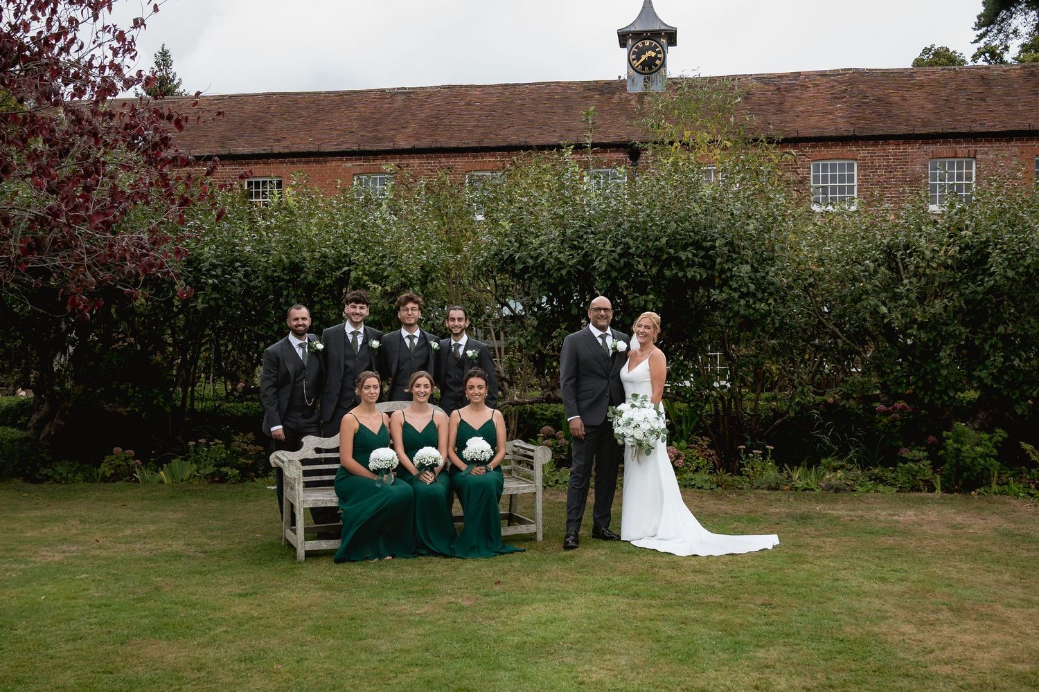 Wedding party gathered on lawn with rustic brick building and gardens in background.