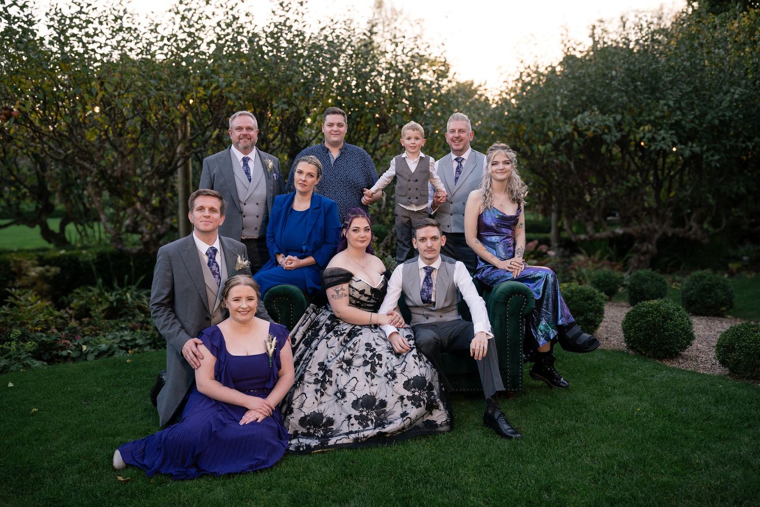 Wedding party gathered on lawn during sunset for group photos.