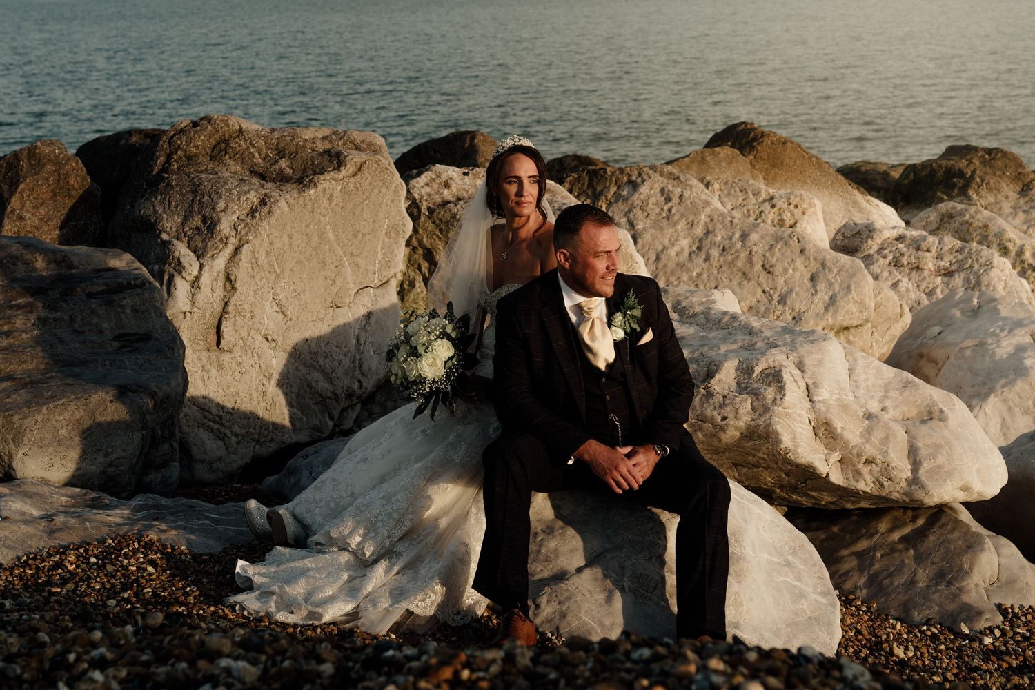 Newlyweds pose together against rocky coastal backdrop at sunset.