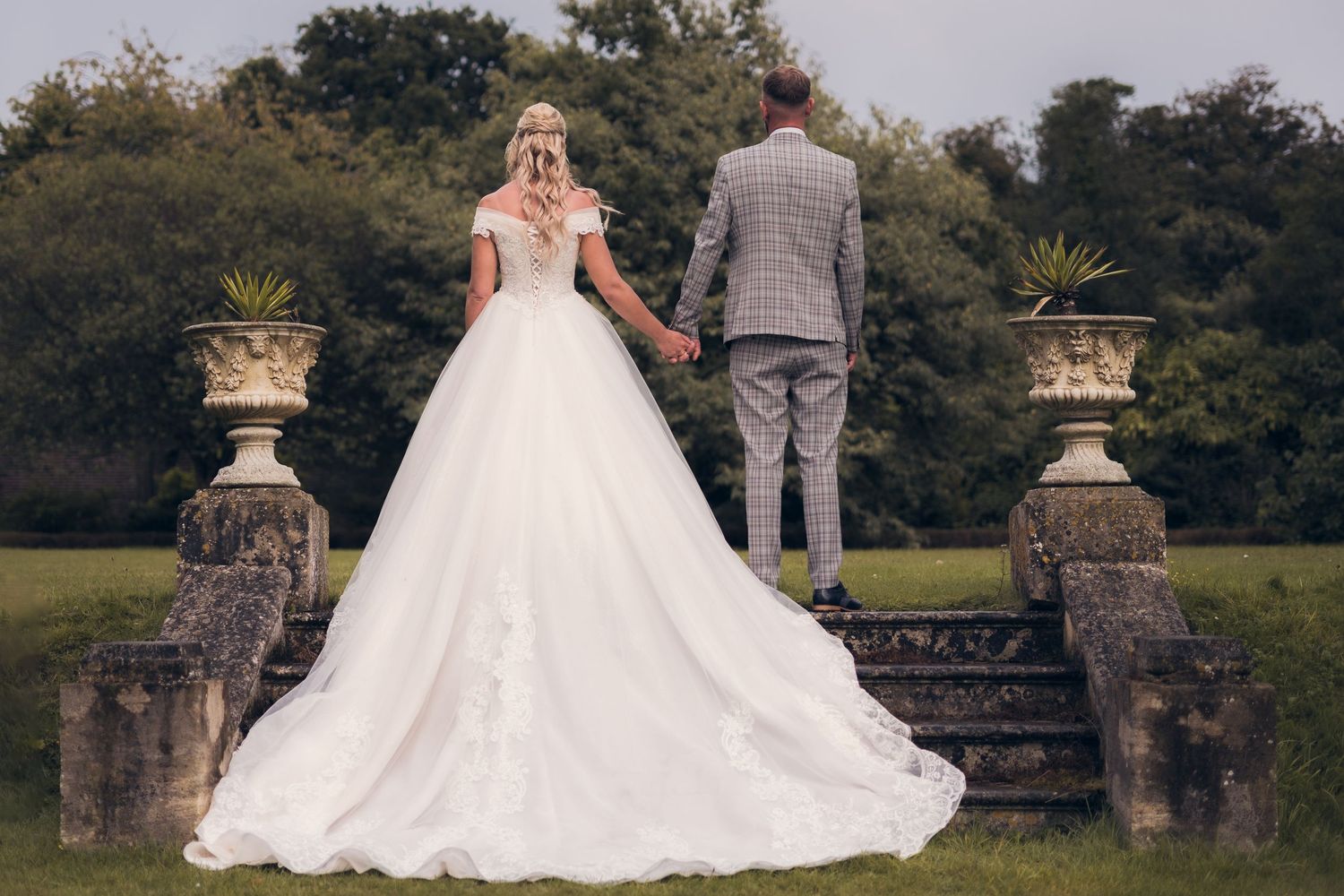 Bride in flowing white gown poses near ornate stone pillars in garden setting.