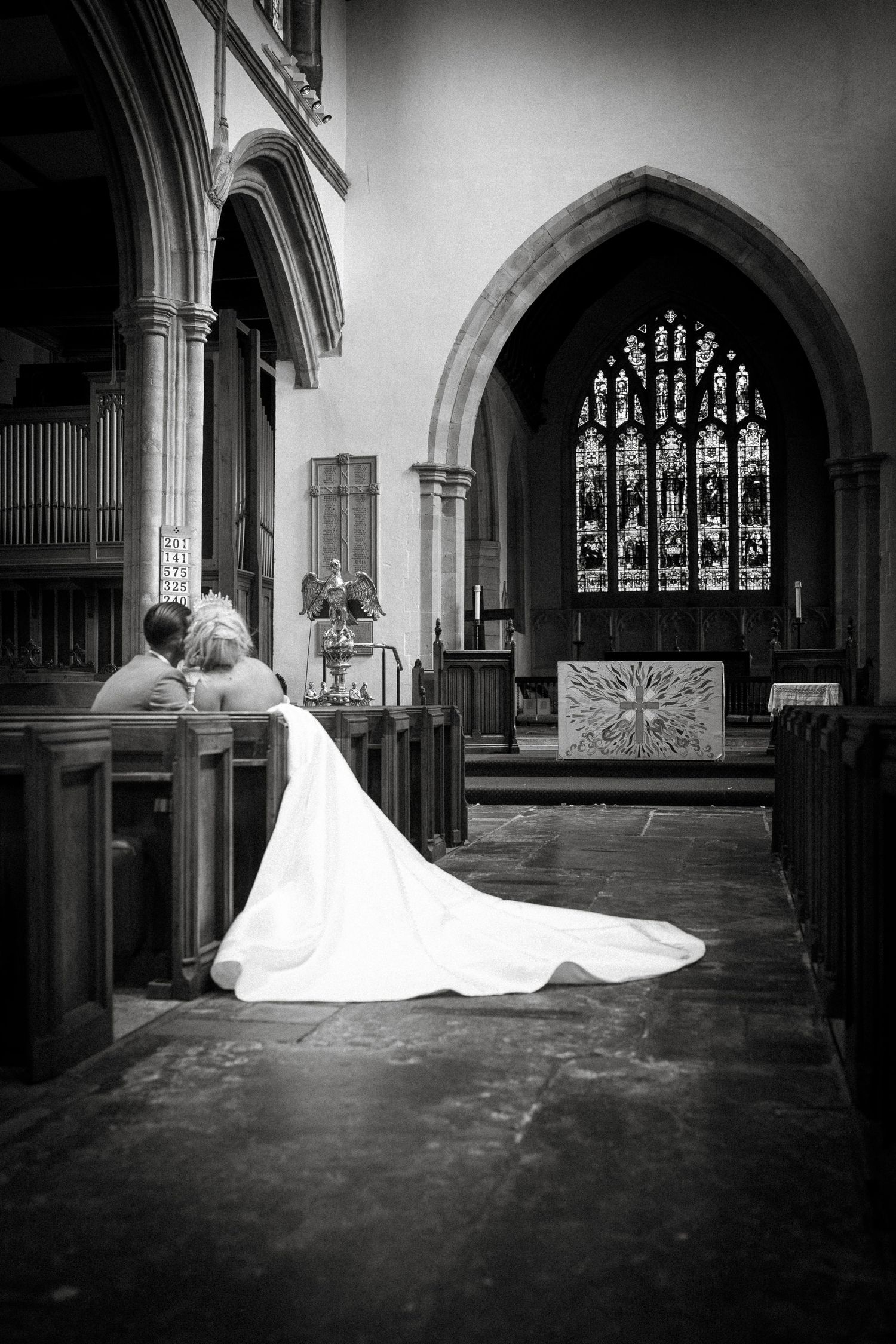 Black and white photograph of a wedding couple in a historic church with stained glass windows and arched architecture.