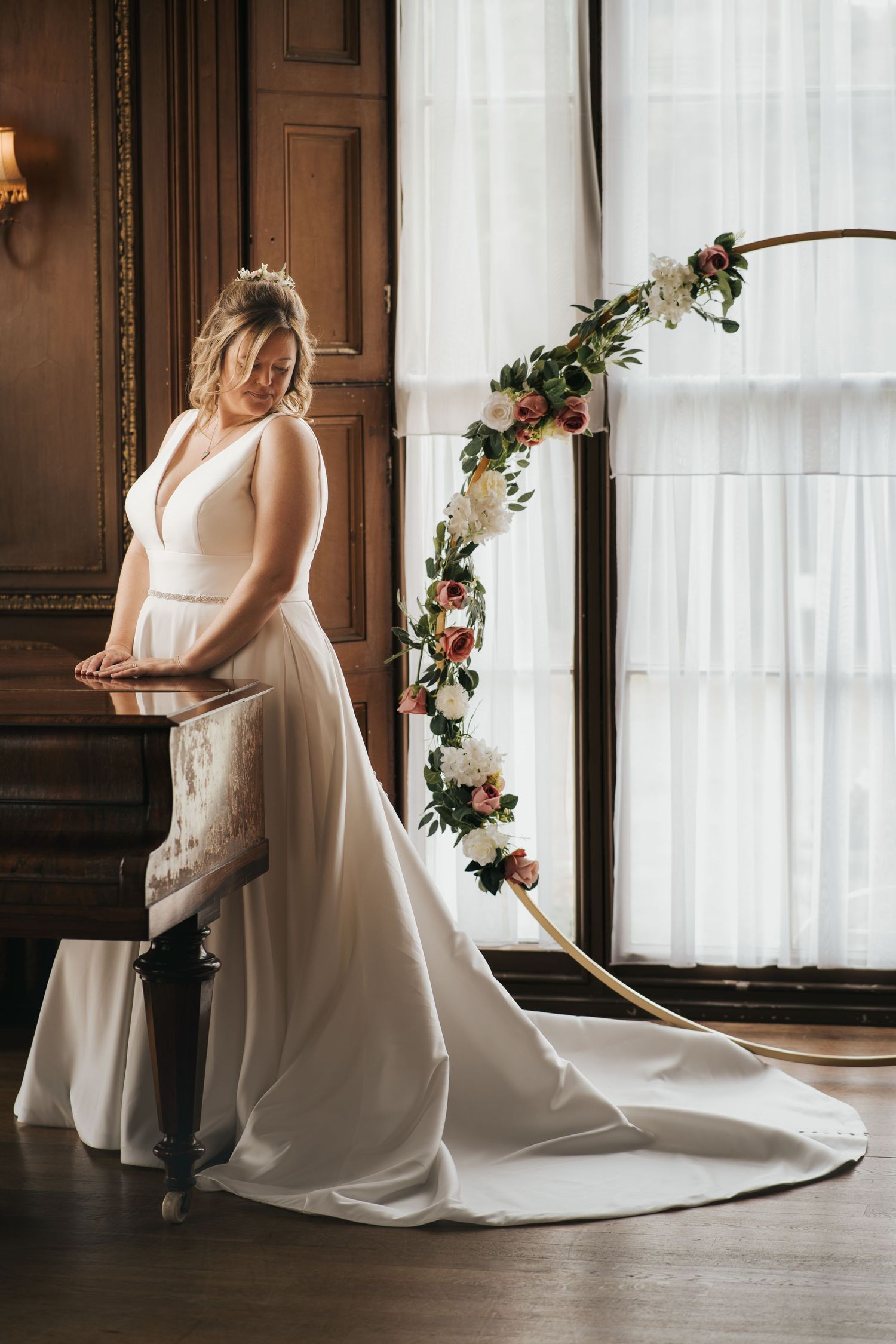 An elegant white wedding dress with a long train flows across a hardwood floor in front of sheer curtains.