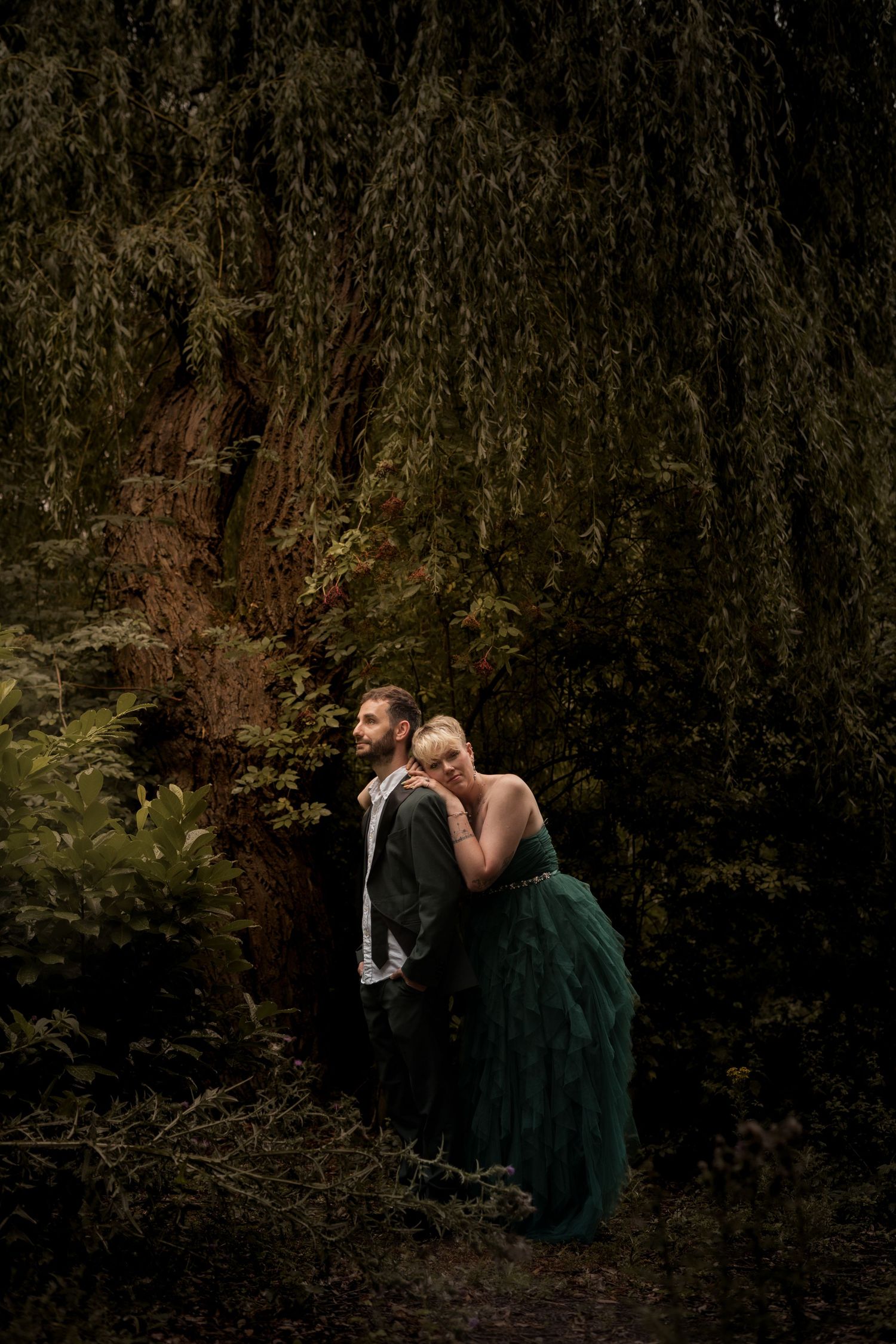 Couple embracing under dark willow branches in a moody forest setting.