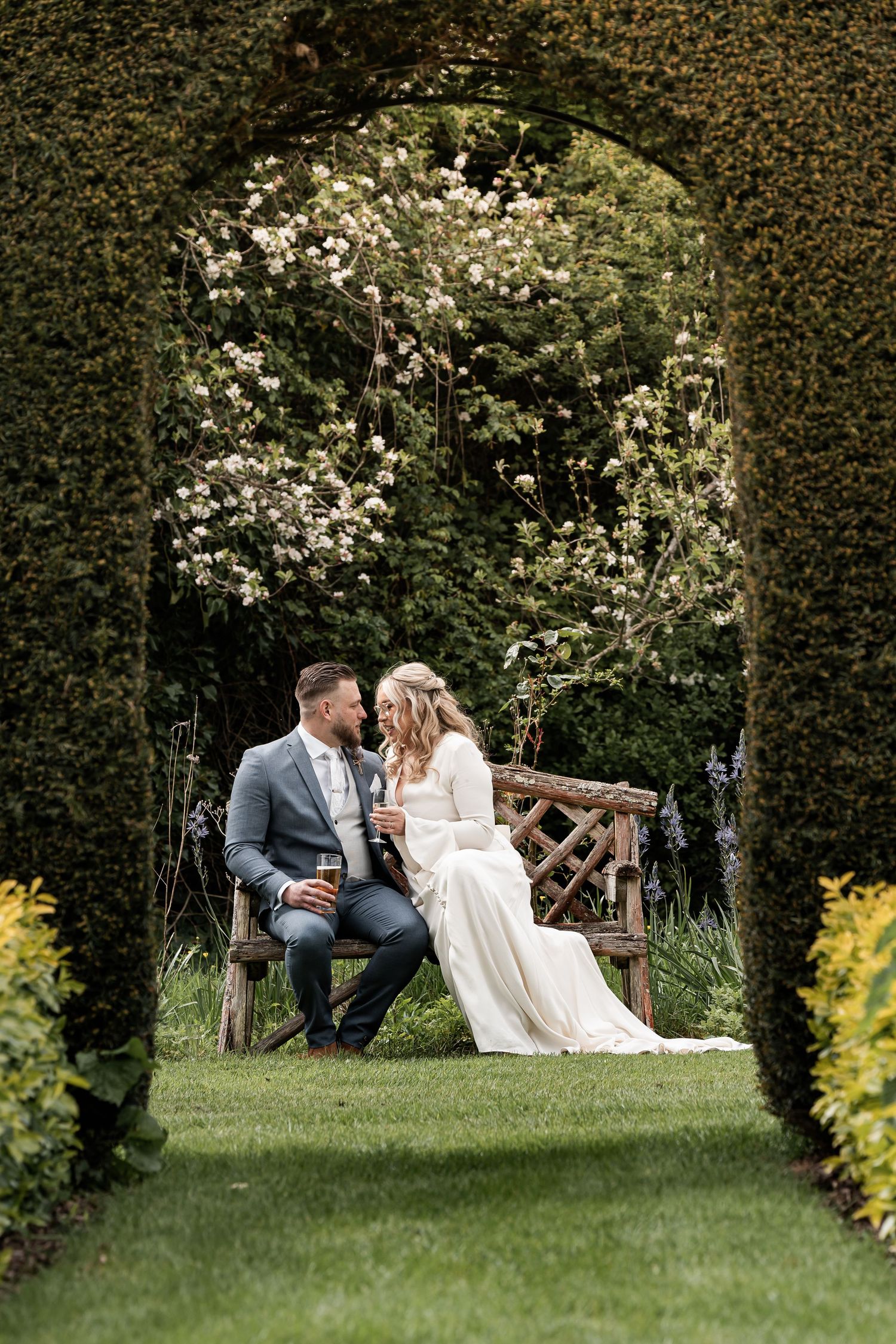 Couple sitting on wooden bench in lush garden setting surrounded by hedges and flowers.