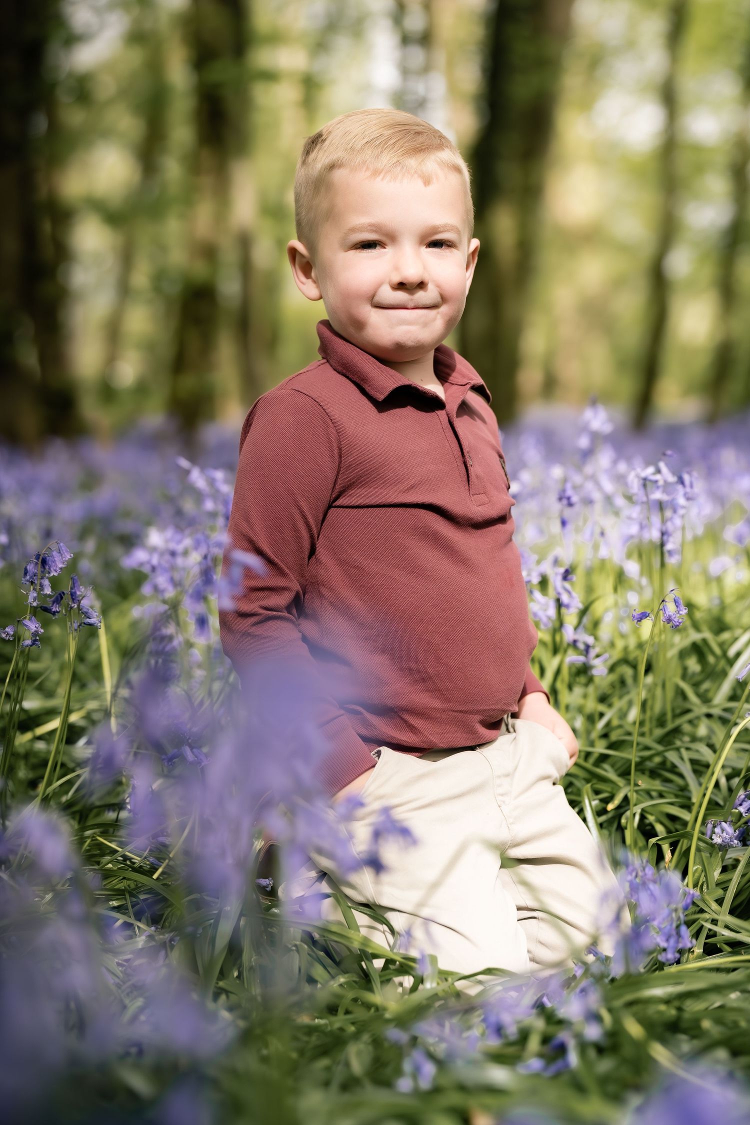 Child wearing brown sweater sits among blooming bluebells in a spring woodland setting.