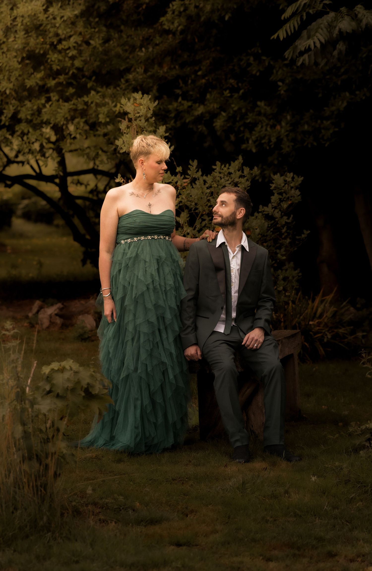 A couple poses together outdoors in formal attire with a green gown and dark suit against a natural backdrop.