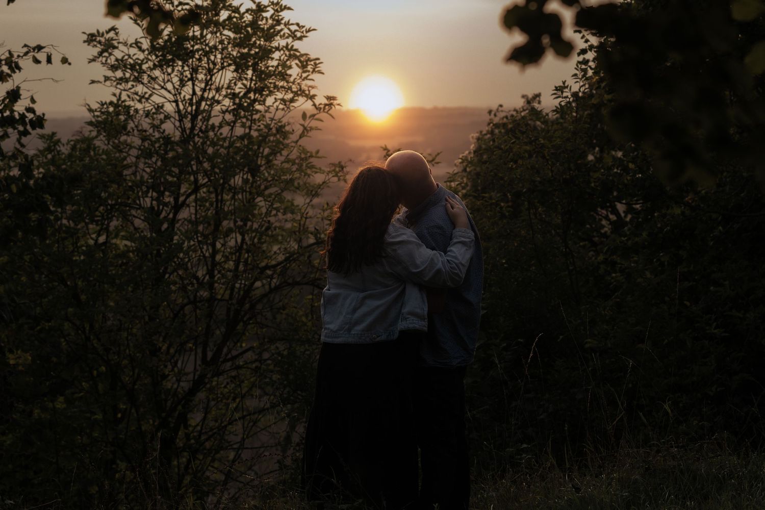 A silhouetted couple embraces against a dramatic sunset sky with trees framing the romantic scene.