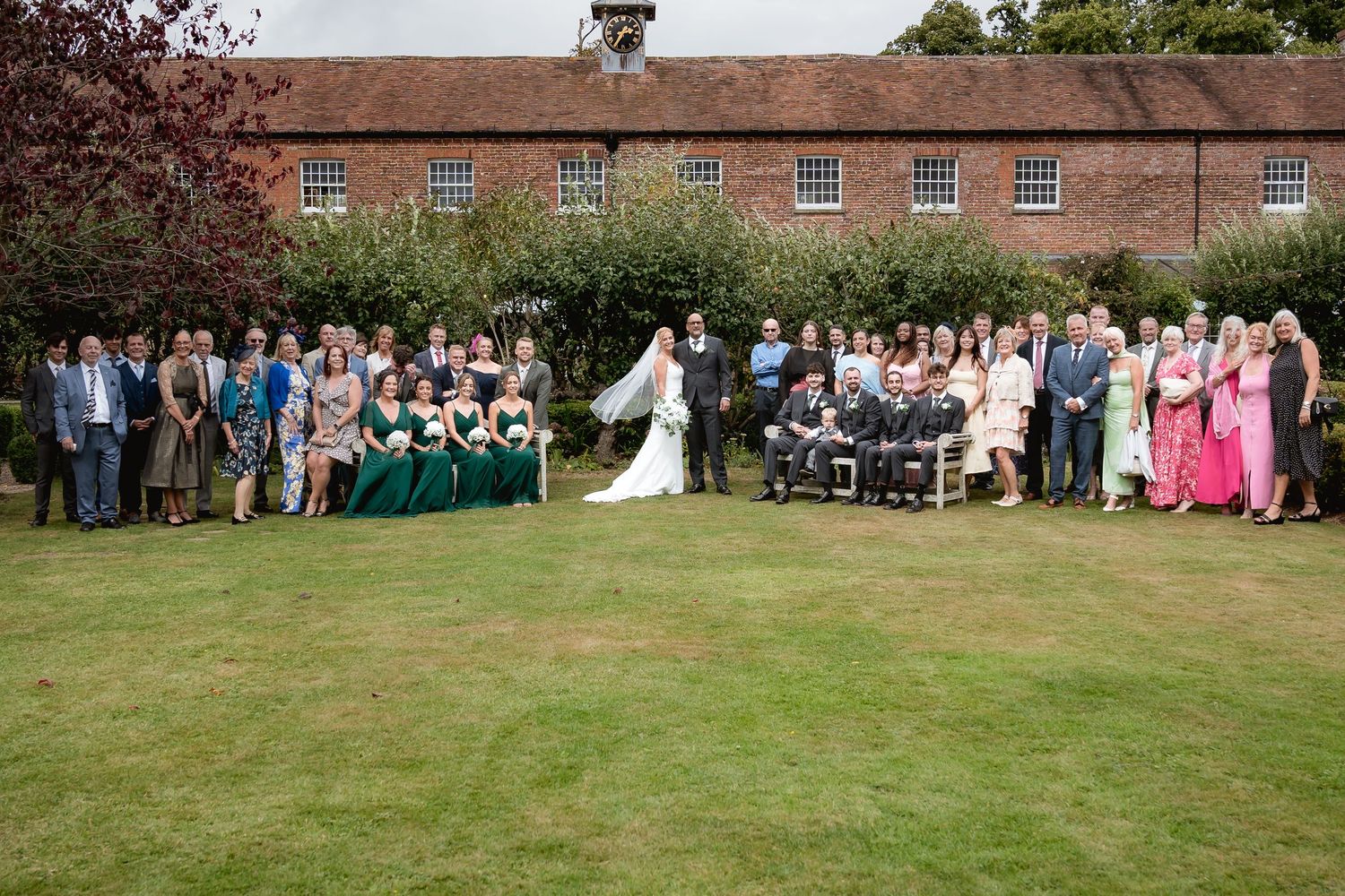Wide panoramic shot of wedding guests seated on a lawn in front of a brick building.