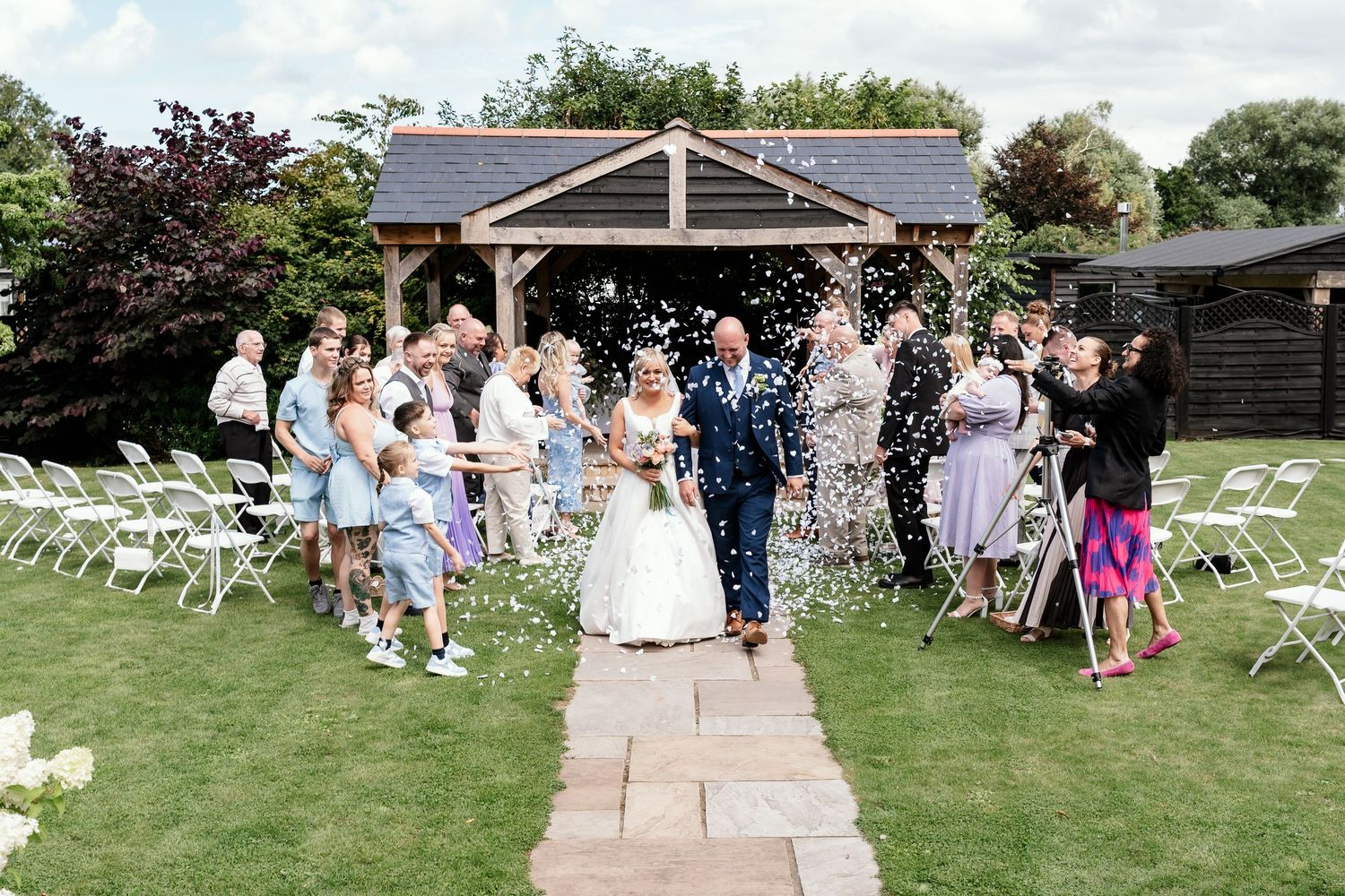 Wedding guests seated in white chairs on a lawn during an outdoor ceremony.