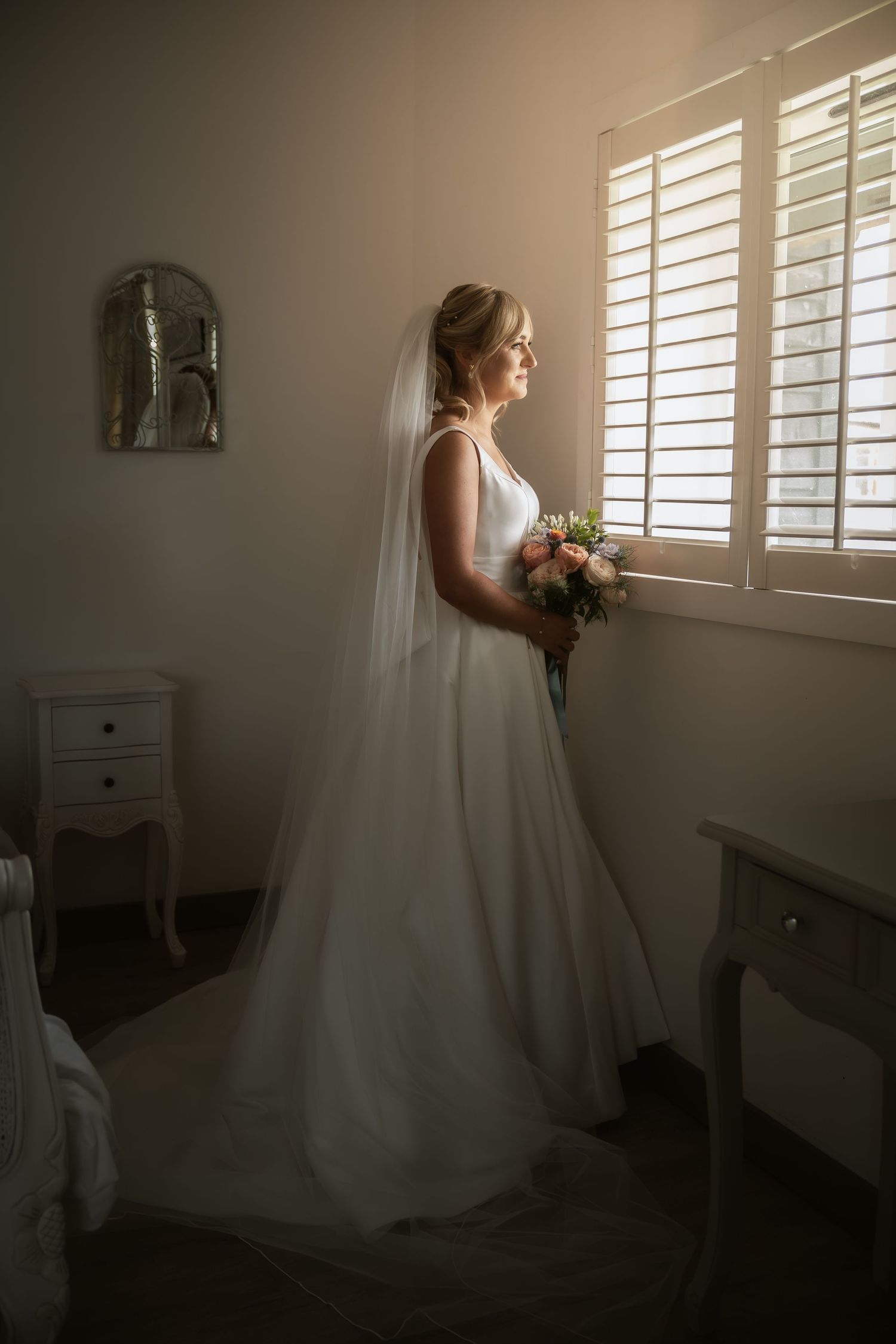 A bride in a flowing white gown stands by a window in a softly lit room.