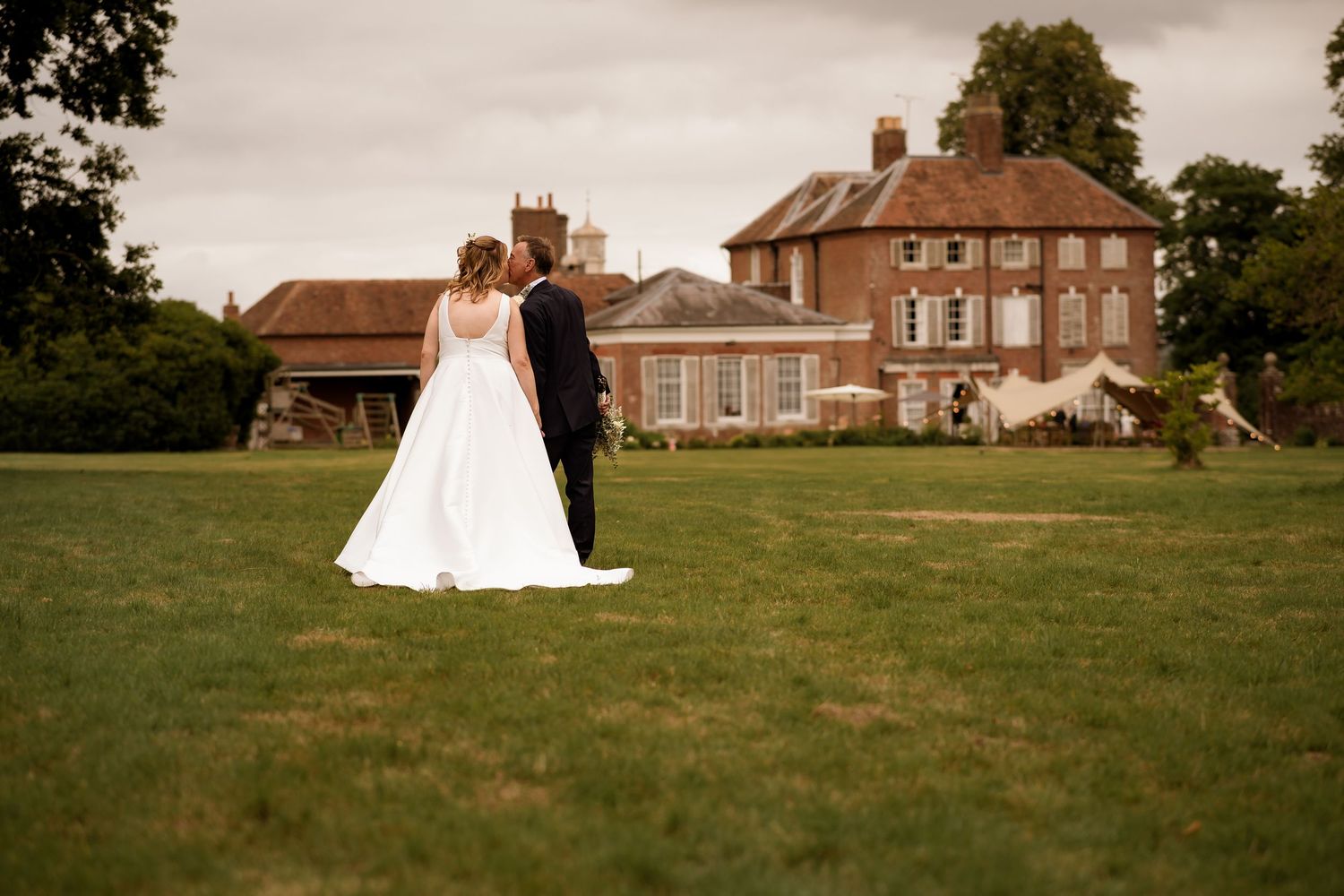 A couple poses on a sprawling green lawn in front of a stately brick mansion on their wedding day.