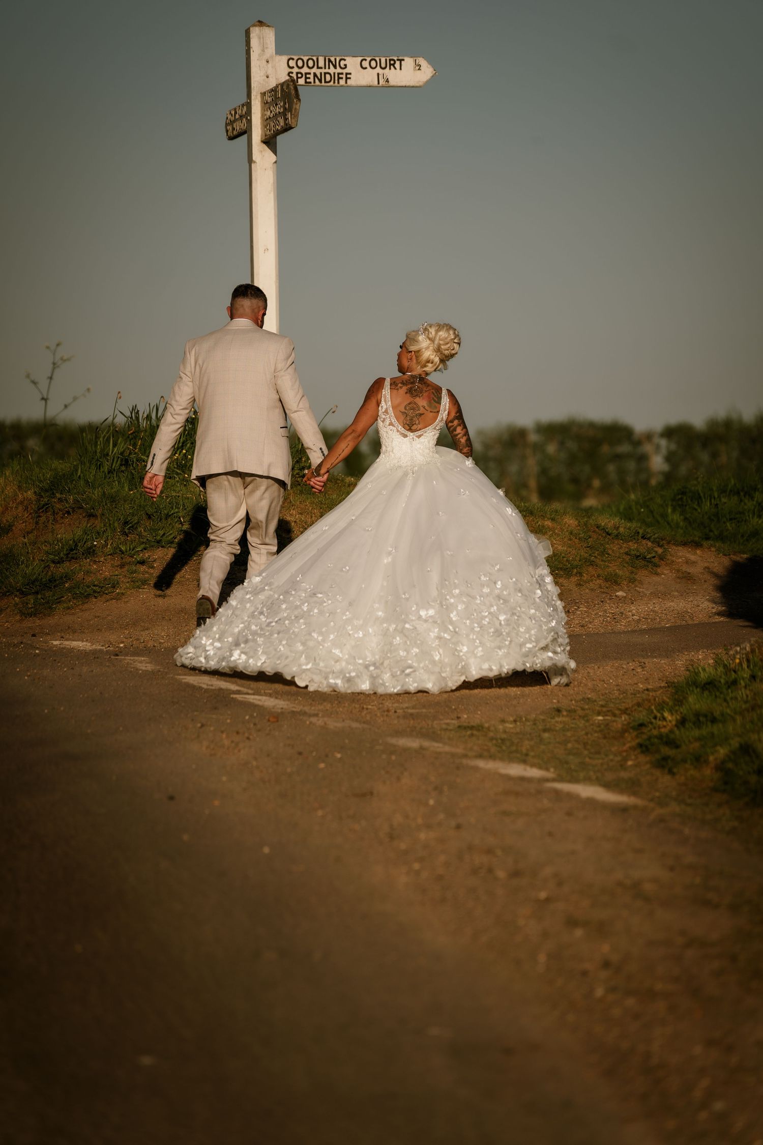 Couple in wedding attire walking hand in hand near countryside signpost at sunset.