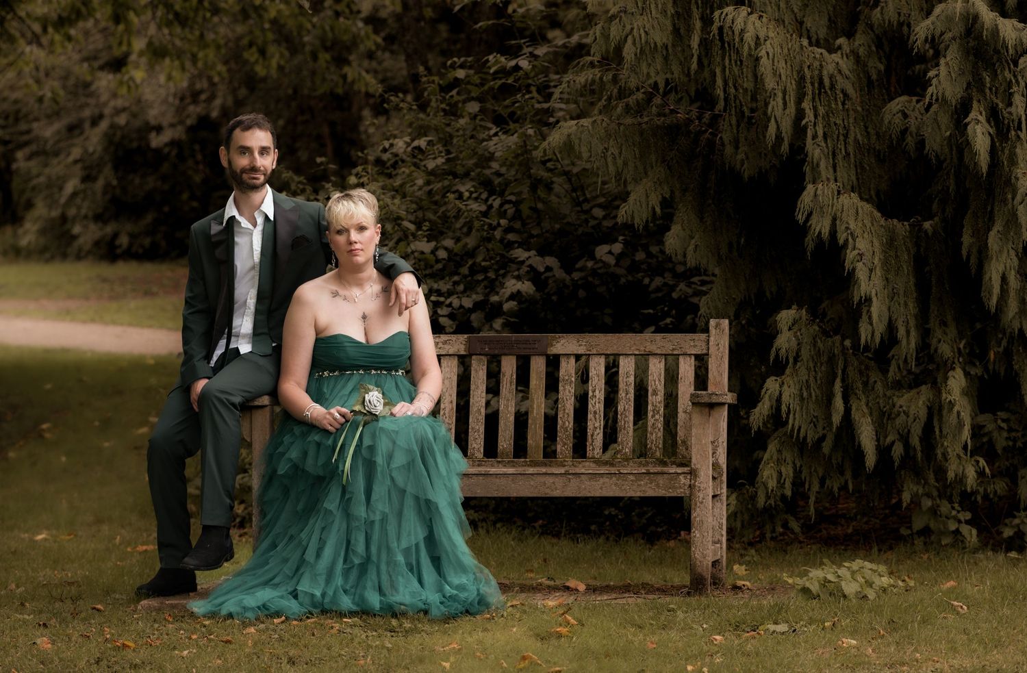 A couple sits together on a wooden bench in formal wear surrounded by garden scenery.