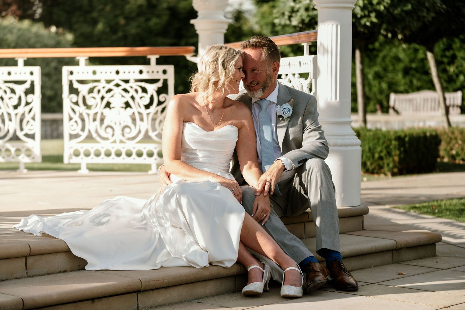 Elegant wedding couple posing on ornate white garden bench in outdoor setting.
