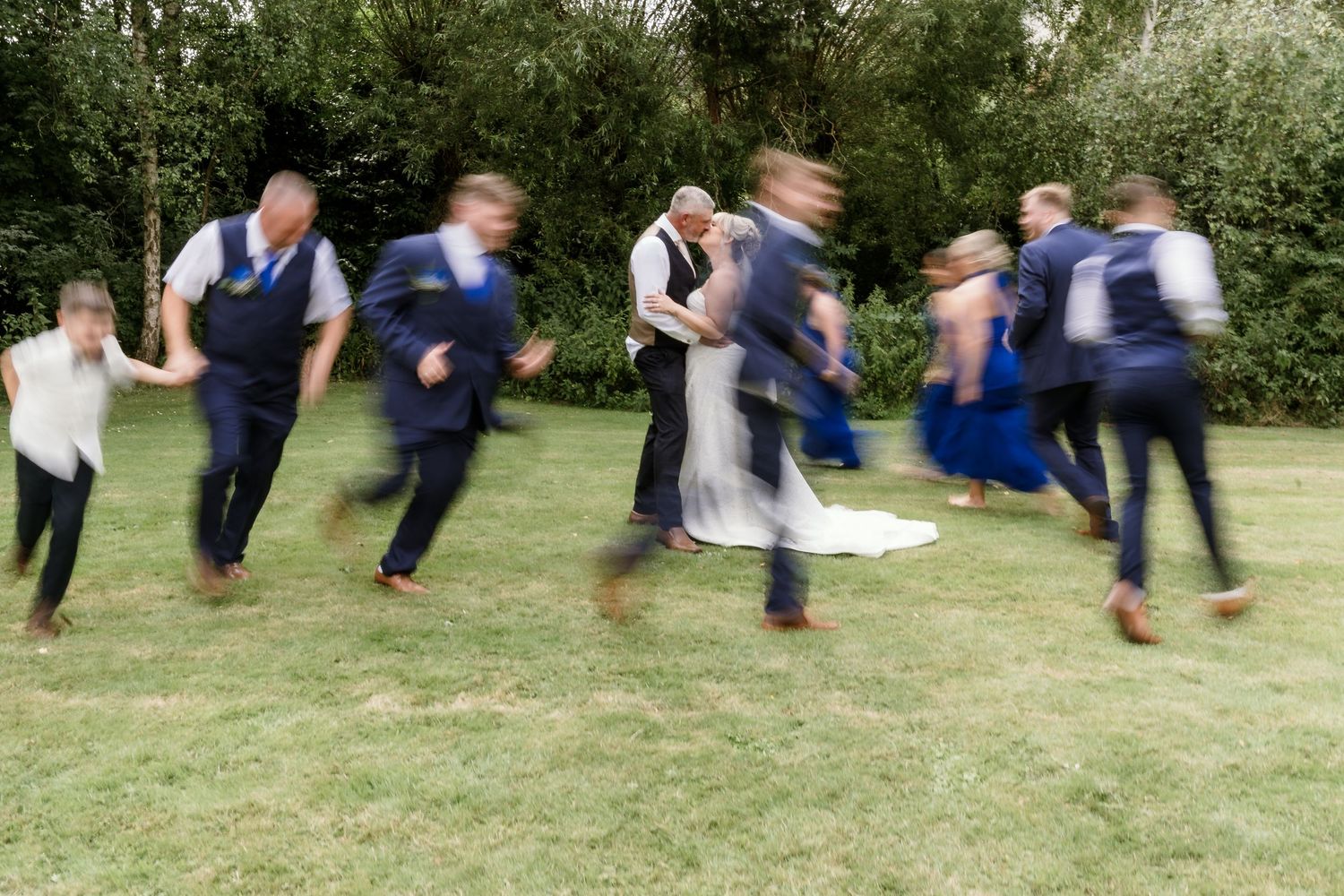 Motion blur photo of wedding guests running and playing on grass lawn during celebration.