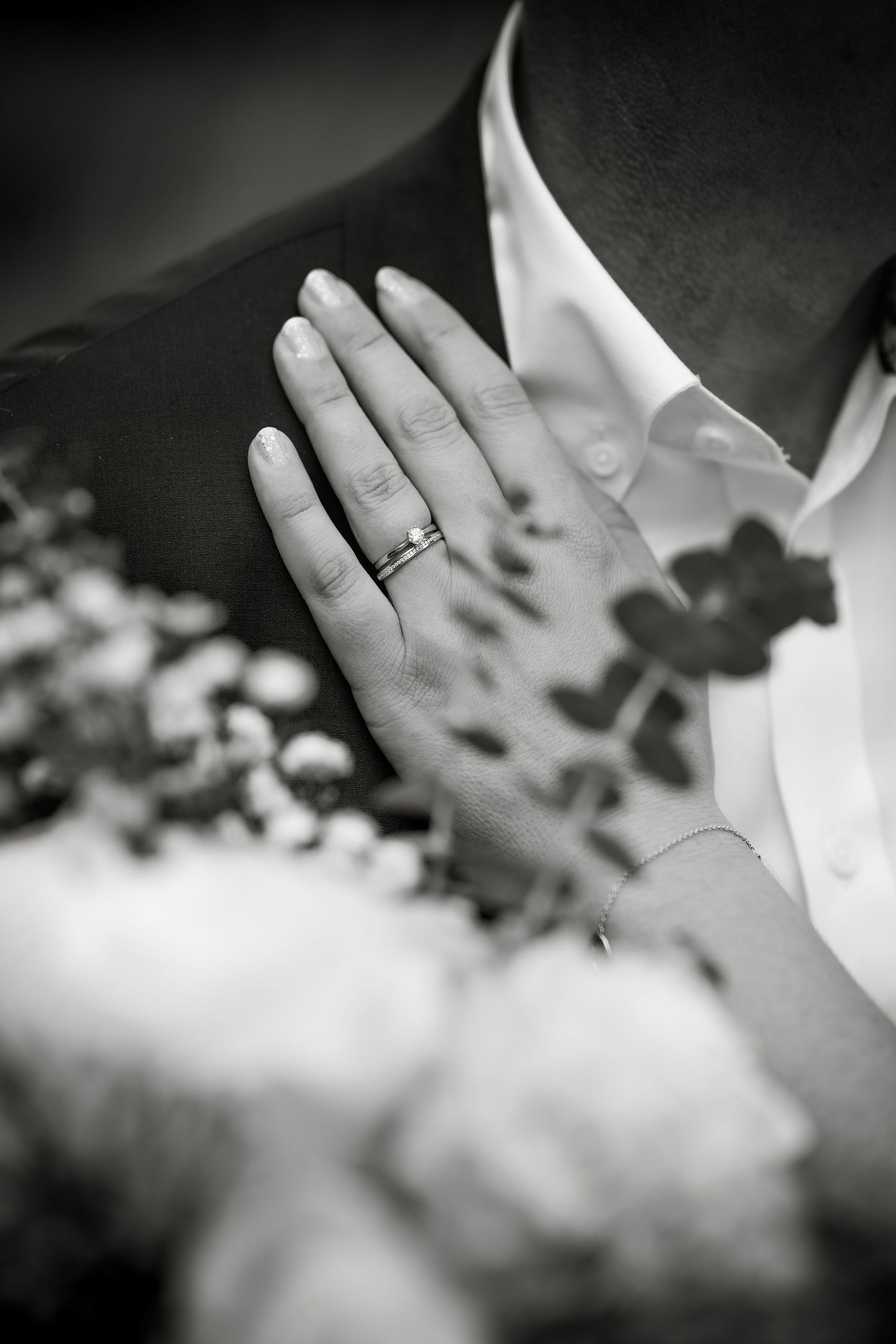 Close-up black and white photo of hands wearing wedding rings with soft floral elements in background.
