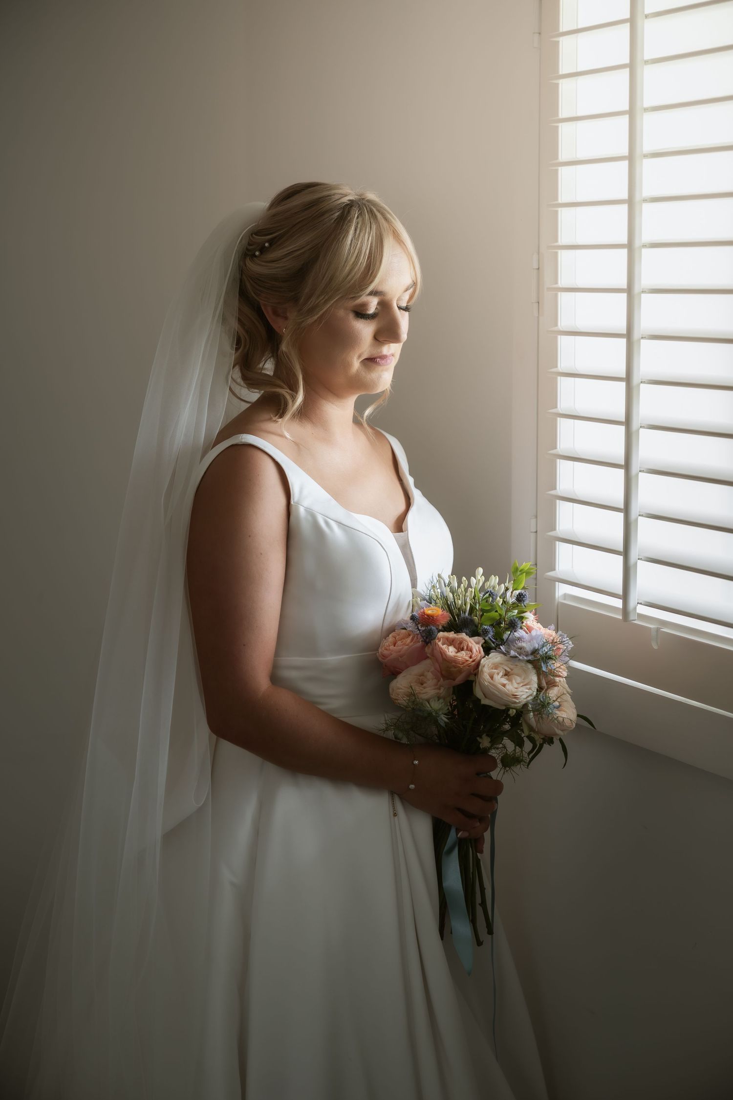 Bride in white dress holds bouquet while standing by window in soft natural light.