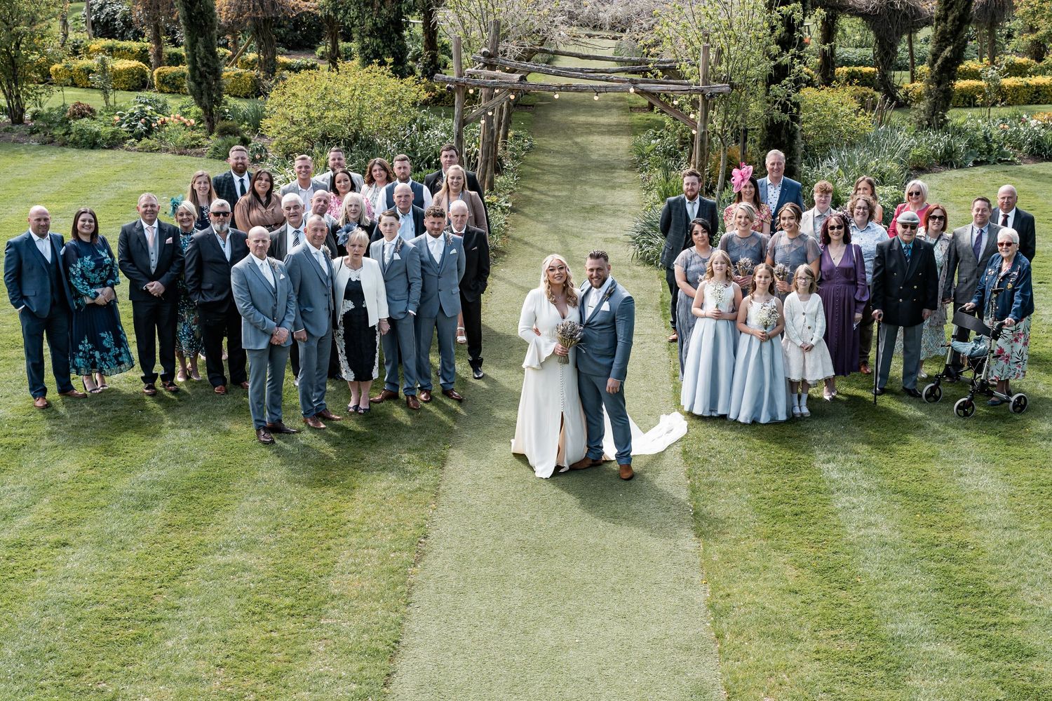 Large wedding party group photo arranged on a sunny green lawn with trees in background.