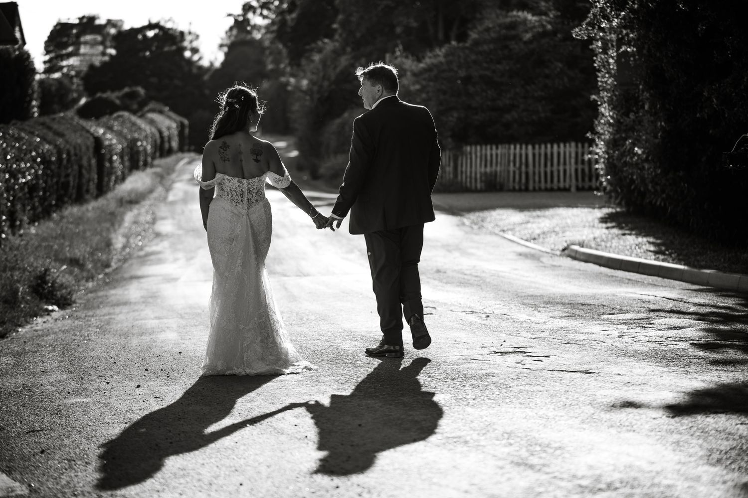 A couple walking down a sunlit path holding hands while casting long shadows in black and white photography.