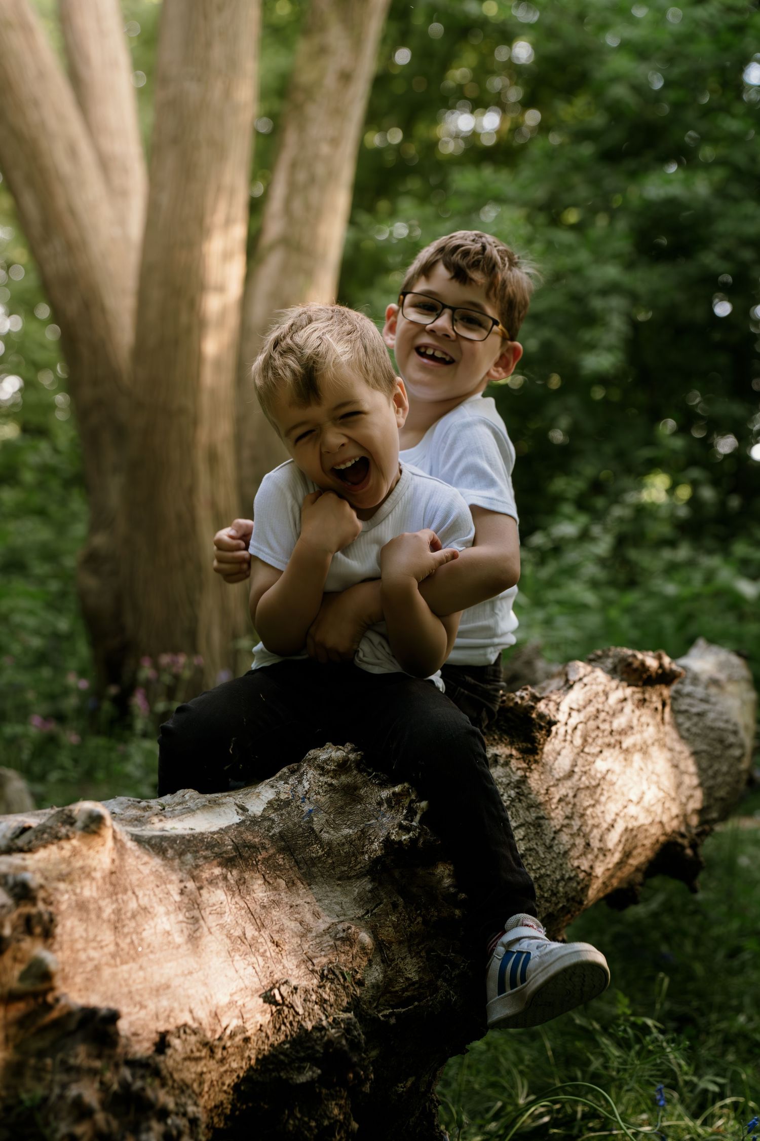 Two children share a joyful moment while sitting together on a fallen log in a wooded outdoor setting.