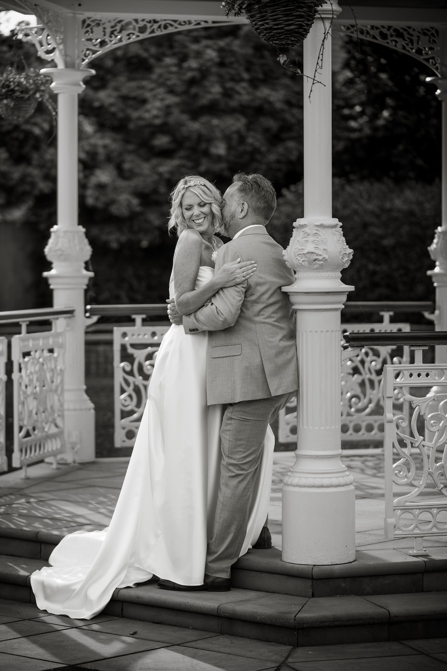Black and white photo of couple sharing tender moment on ornate white gazebo.
