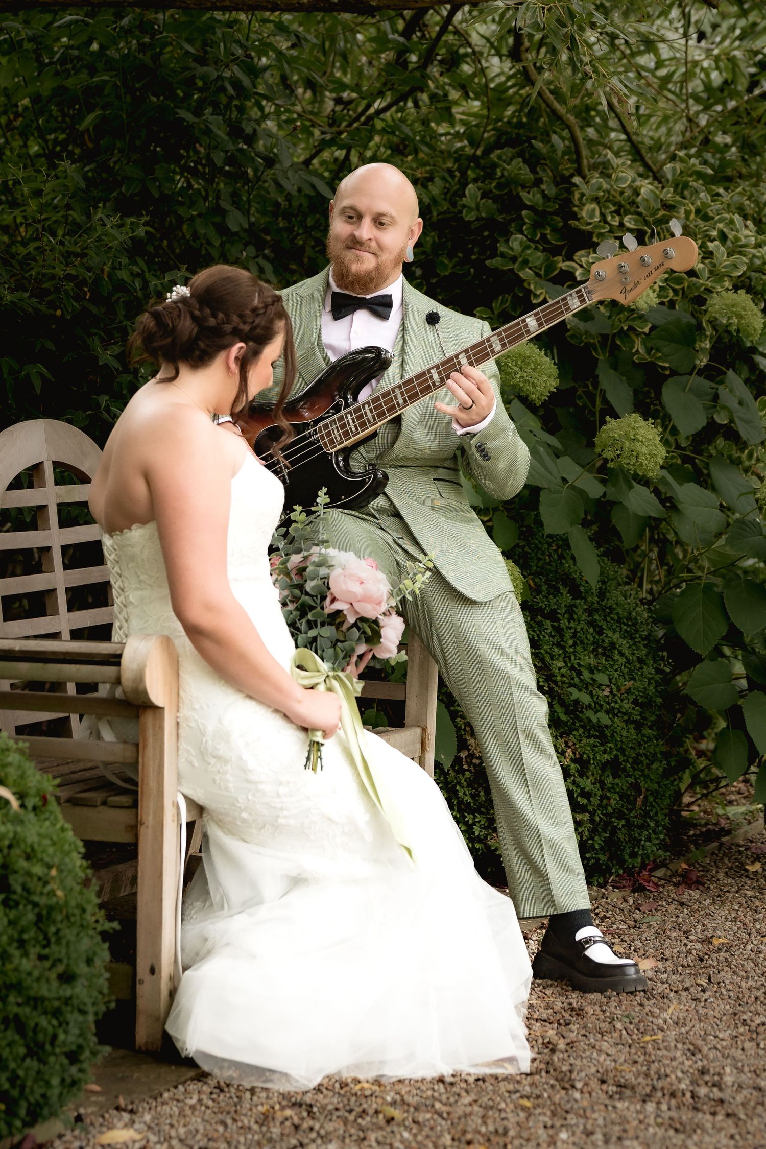 Wedding couple poses near garden bench while groom plays bass guitar.