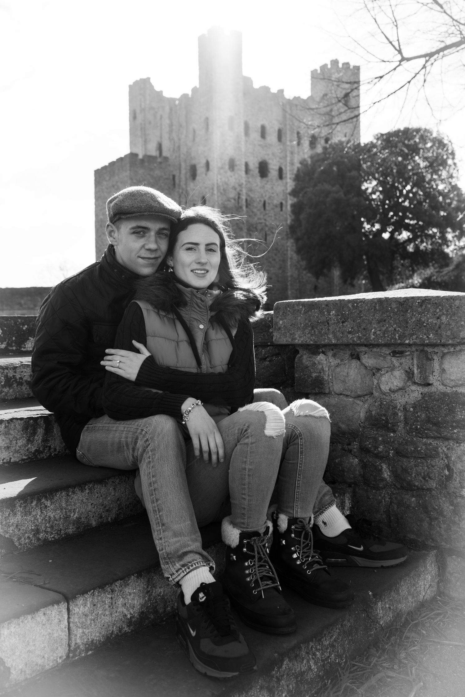 Couple sitting together on stone wall with castle backdrop.