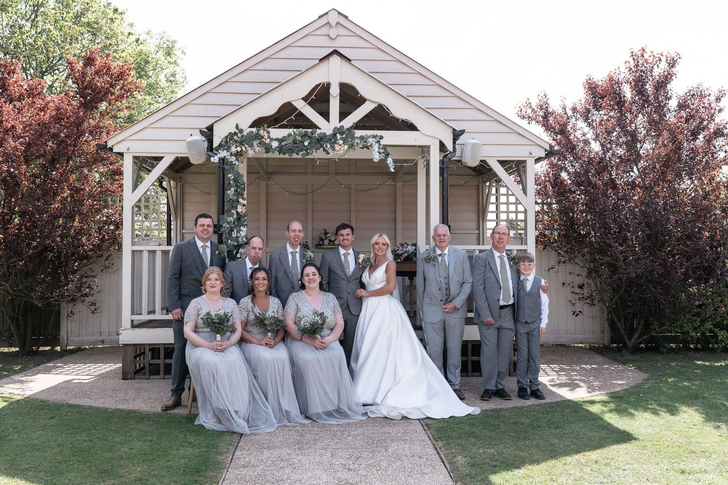 Wedding party in grey and white poses in front of rustic gazebo.