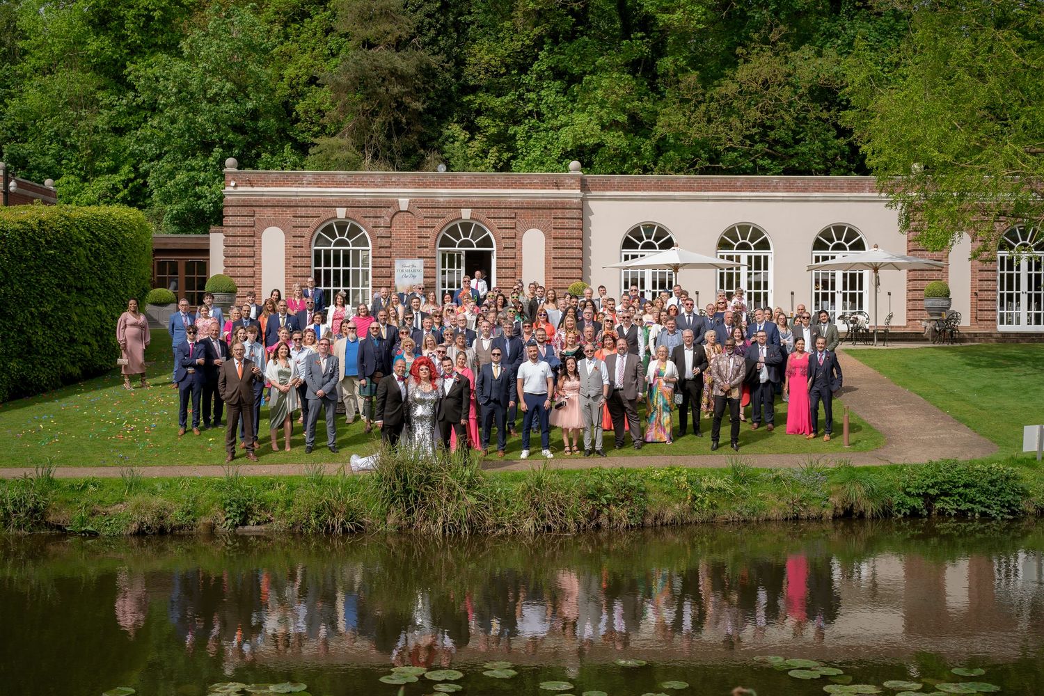 Large group of wedding guests pose for a panoramic photo by a reflective pond.