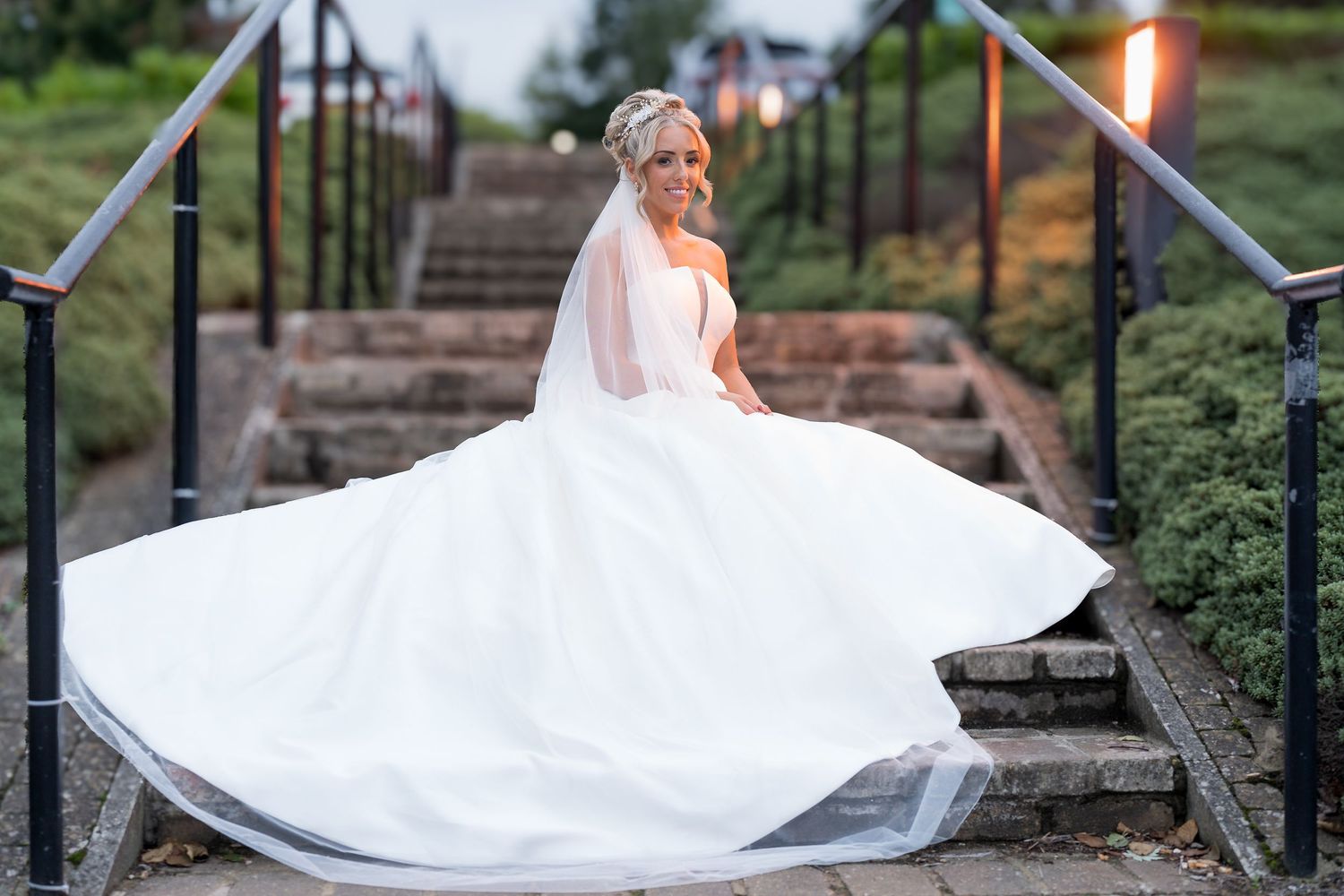A bride in a flowing white dress poses dramatically on stone steps in a garden setting.