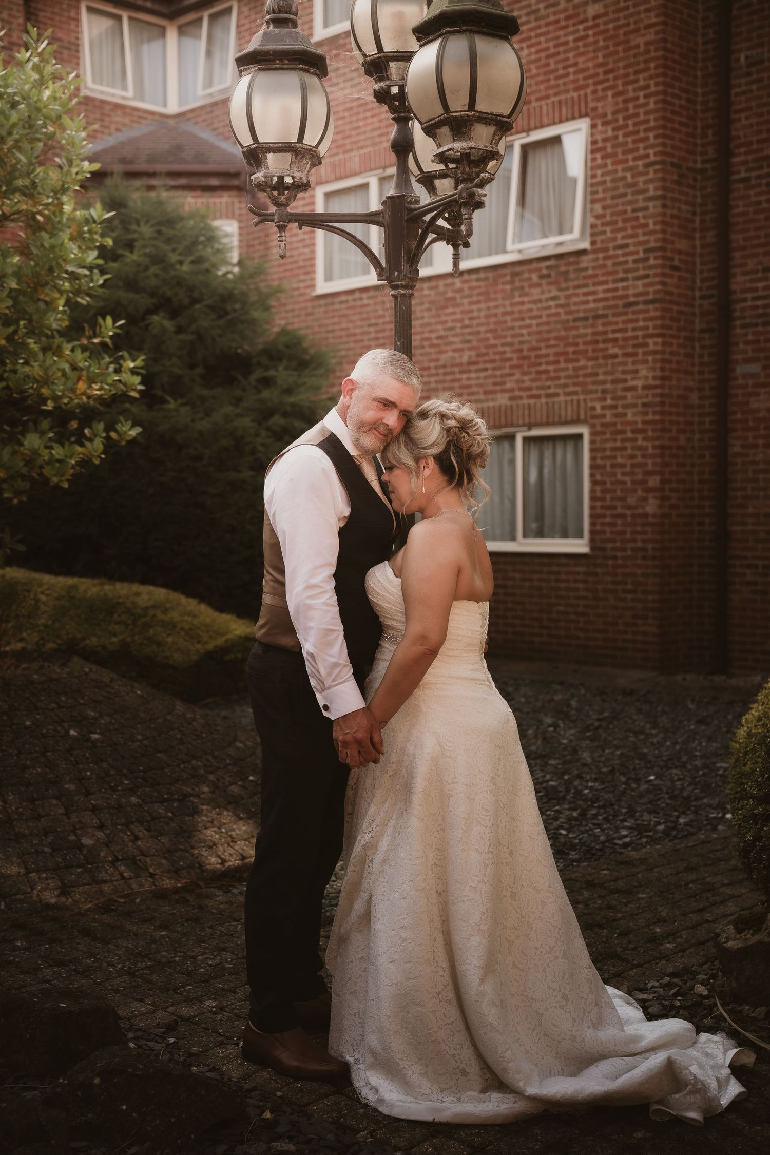 A romantic wedding couple shares an intimate moment against a brick building backdrop at sunset.