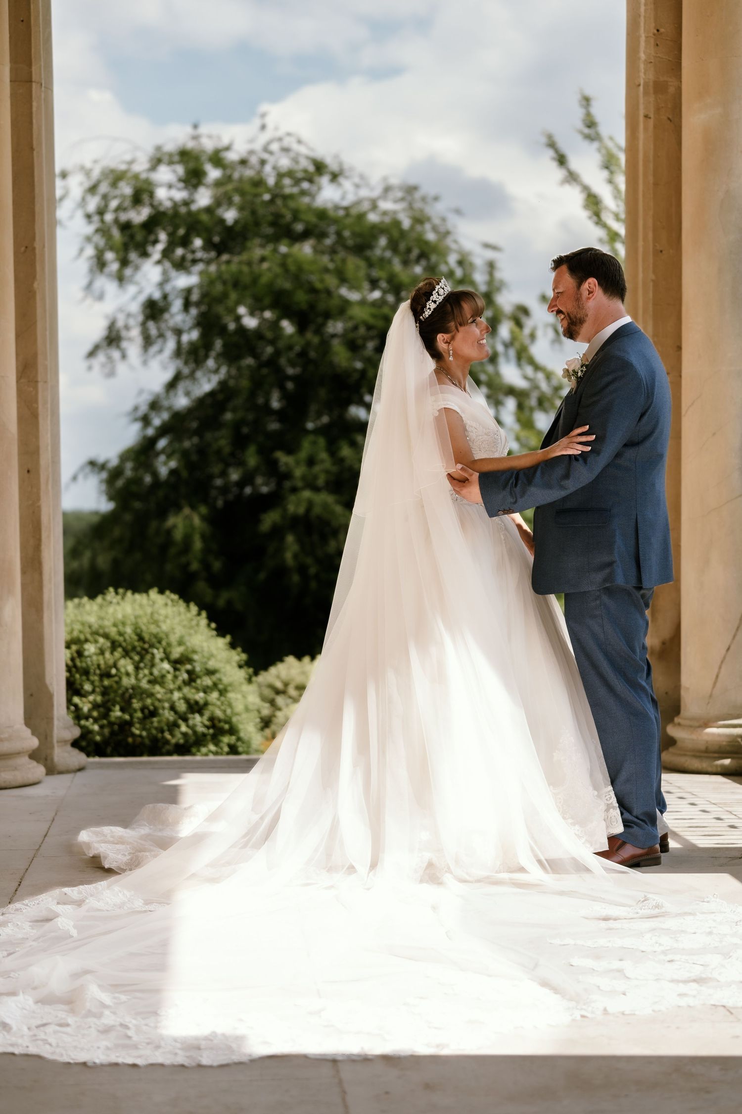 Wedding couple shares a tender moment under classical stone columns outdoors.