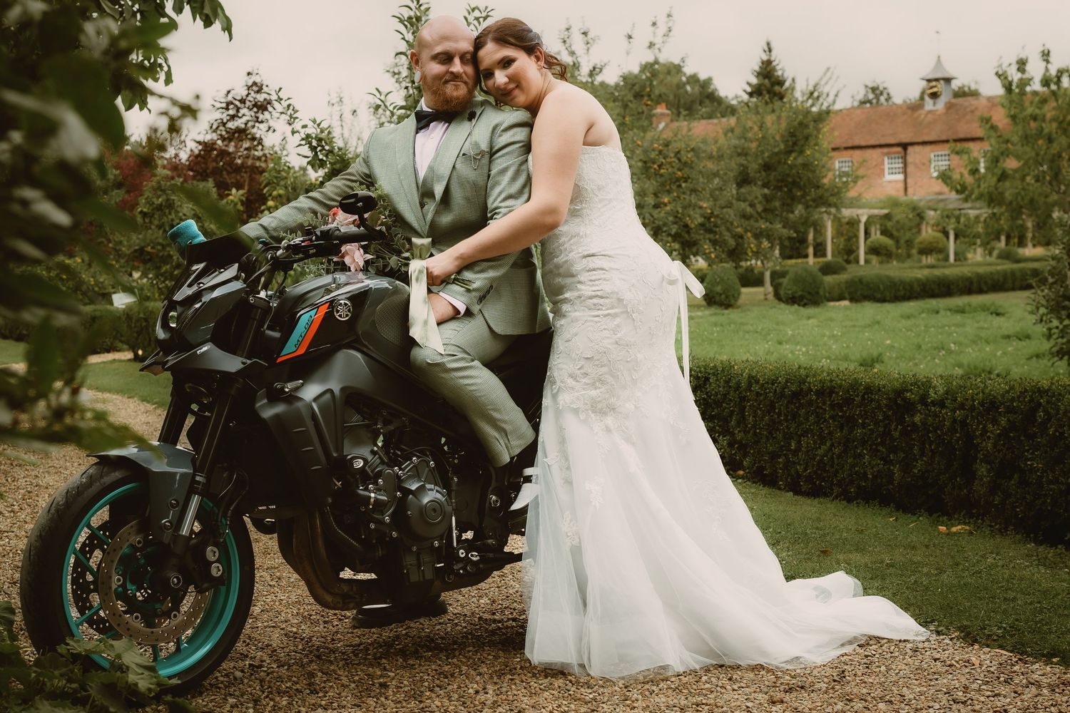 Bride and groom posing with vintage motorcycle in garden setting.