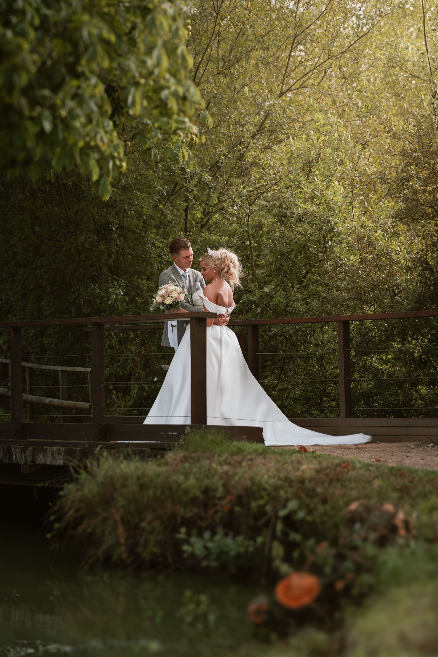Two people in white dresses embracing on a rustic bridge surrounded by lush greenery.