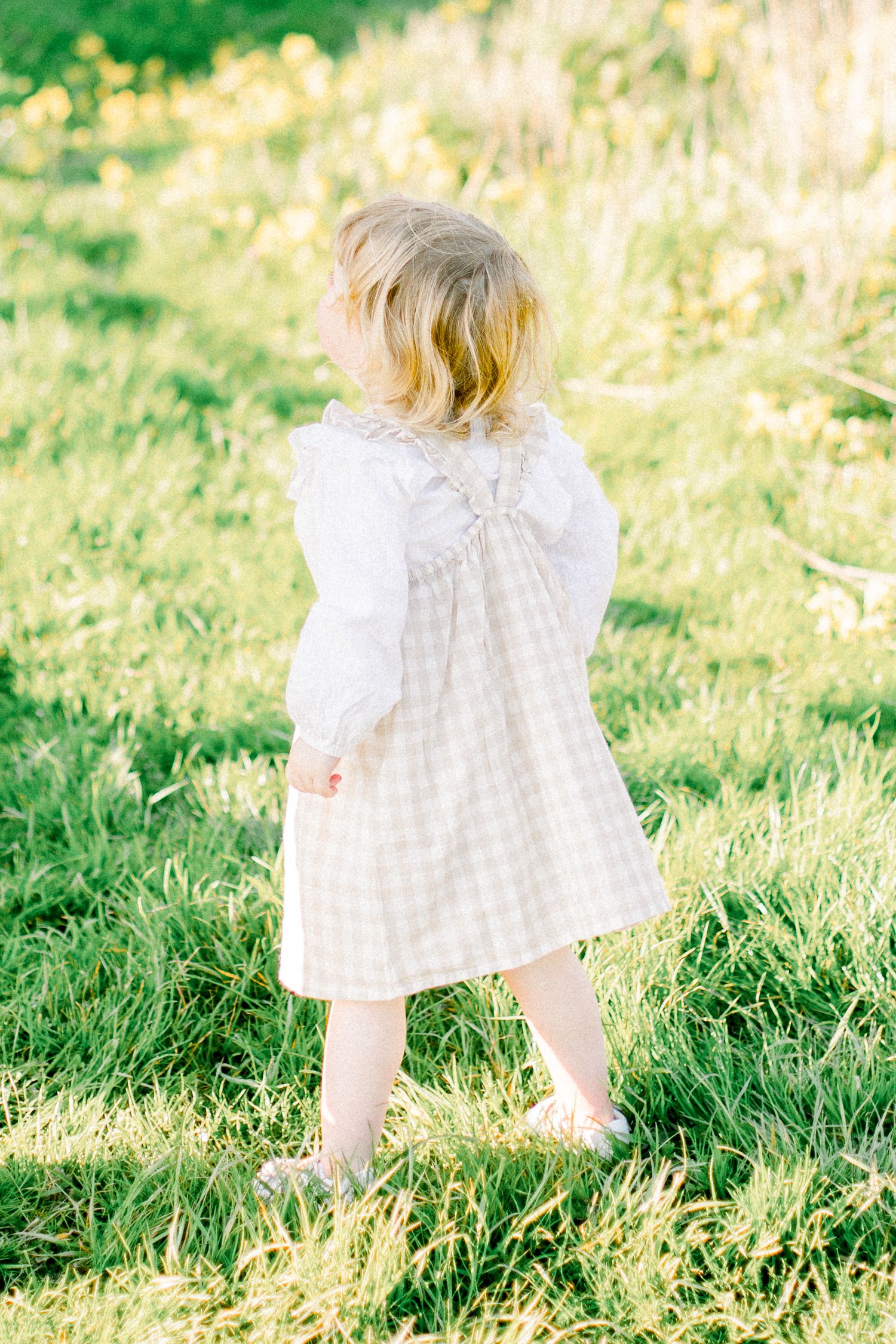 A figure in a white checkered dress walking through sunlit grass during golden hour.