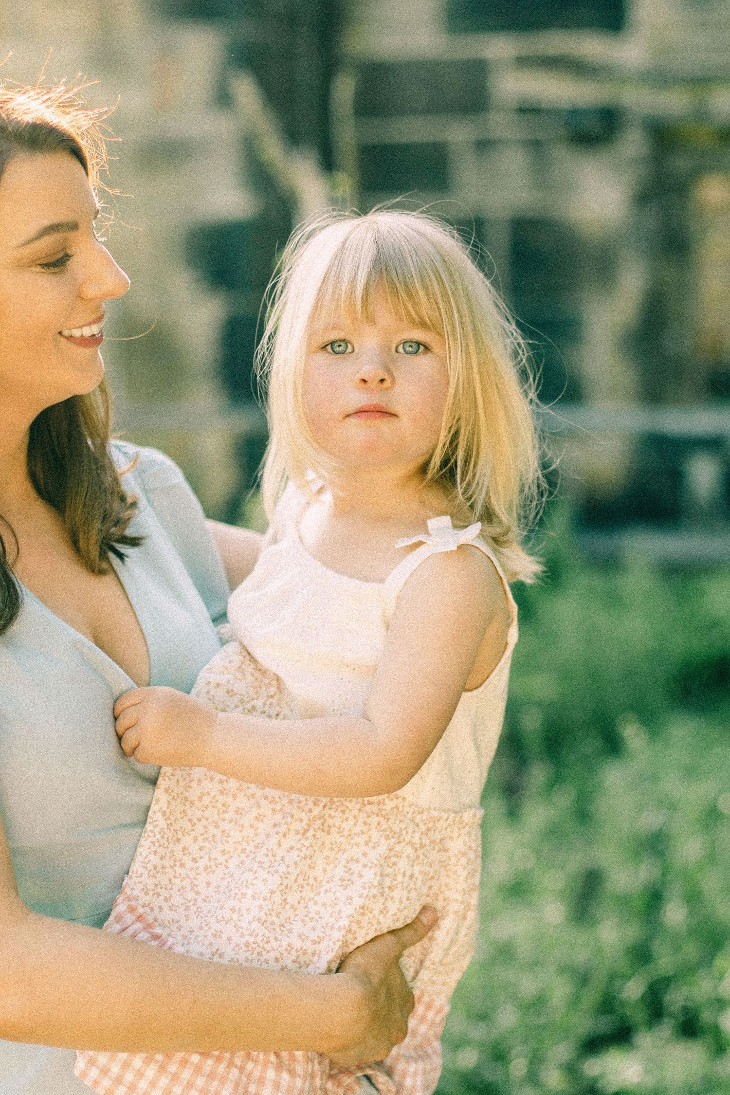 Smiling woman in a light blue top holds a blonde toddler in a pink floral dress outdoors, with green foliage and a stone wall behind them.