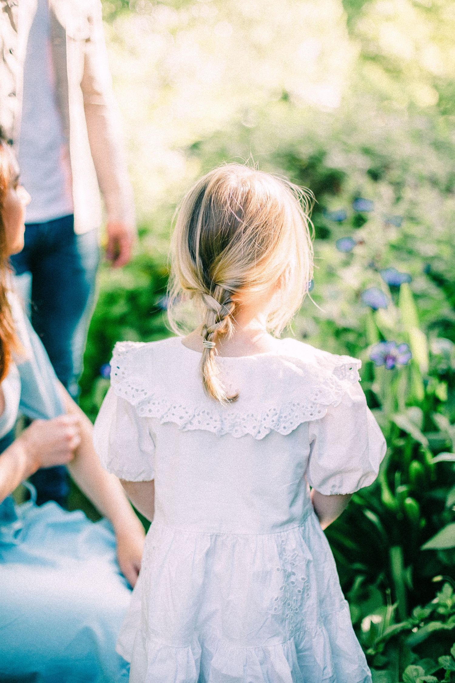 Little girl with a loose blonde braid in a white lace-trimmed dress stands facing a sunlit garden of blue flowers beside two adults.