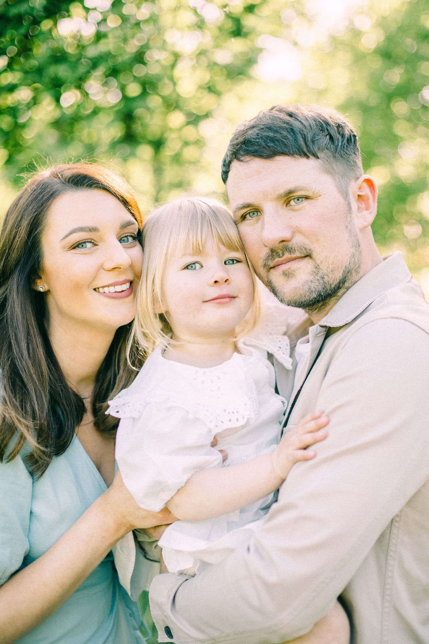 Smiling woman and bearded man in soft neutral outfits hold a blonde toddler in a white lace top outdoors among sunlit green trees.