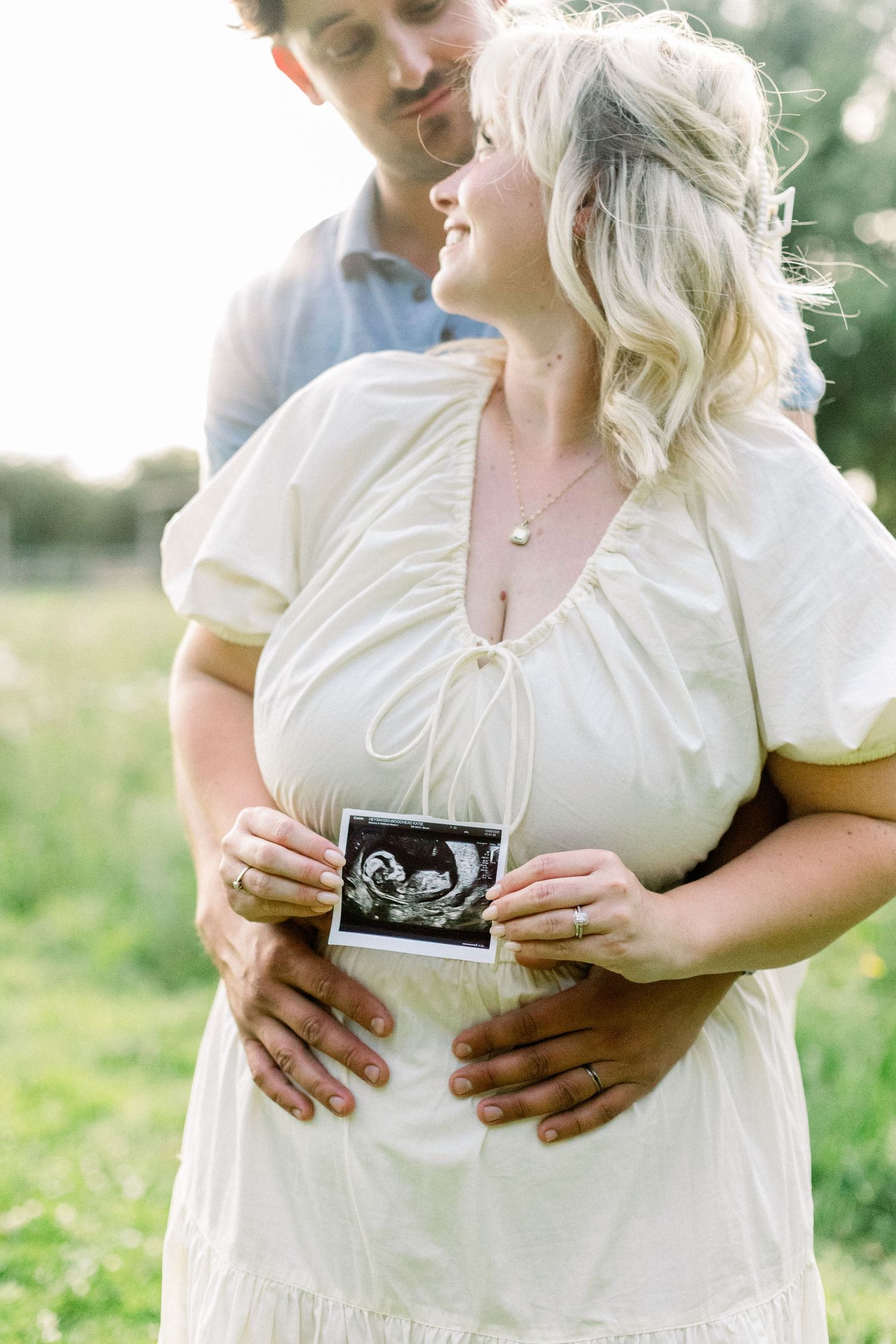 Expectant mother in a white dress holds an ultrasound photo as her partner hugs her belly in a sunlit green field.