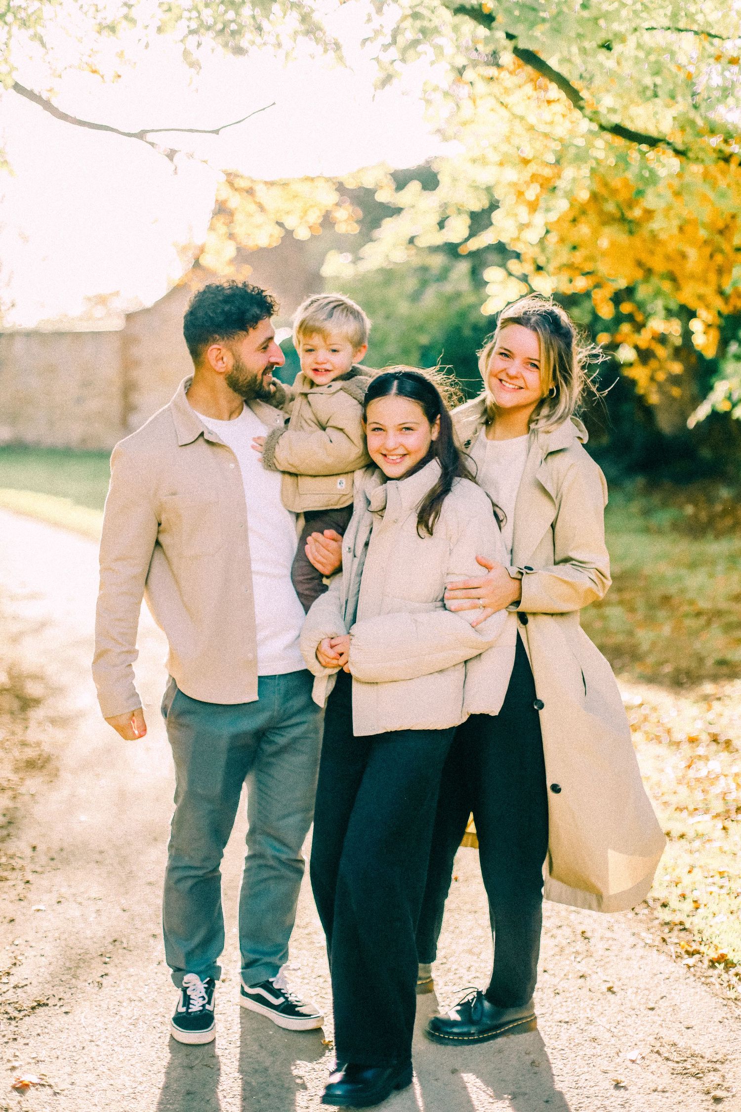 Smiling family of four in matching beige and neutral outfits poses together on a sunlit autumn path lined with golden trees.