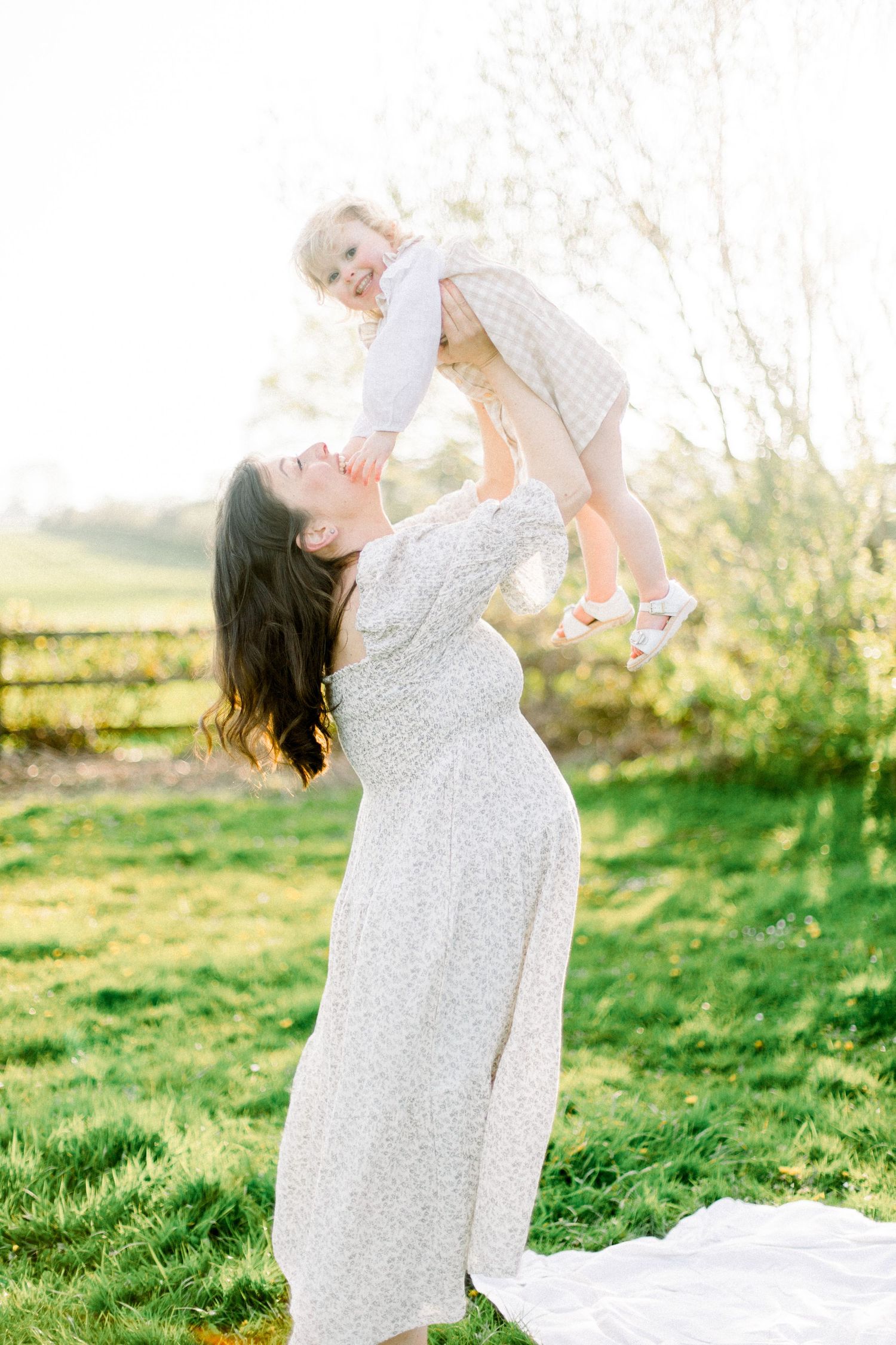 A joyful mother in a floral dress lifts her laughing toddler daughter high in the air in a sunny green garden.