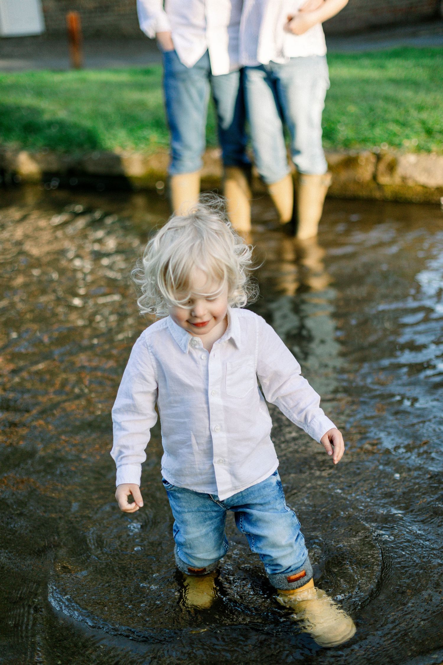 Toddler with blonde curly hair splashing in a muddy puddle wearing rubber boots while parents watch.