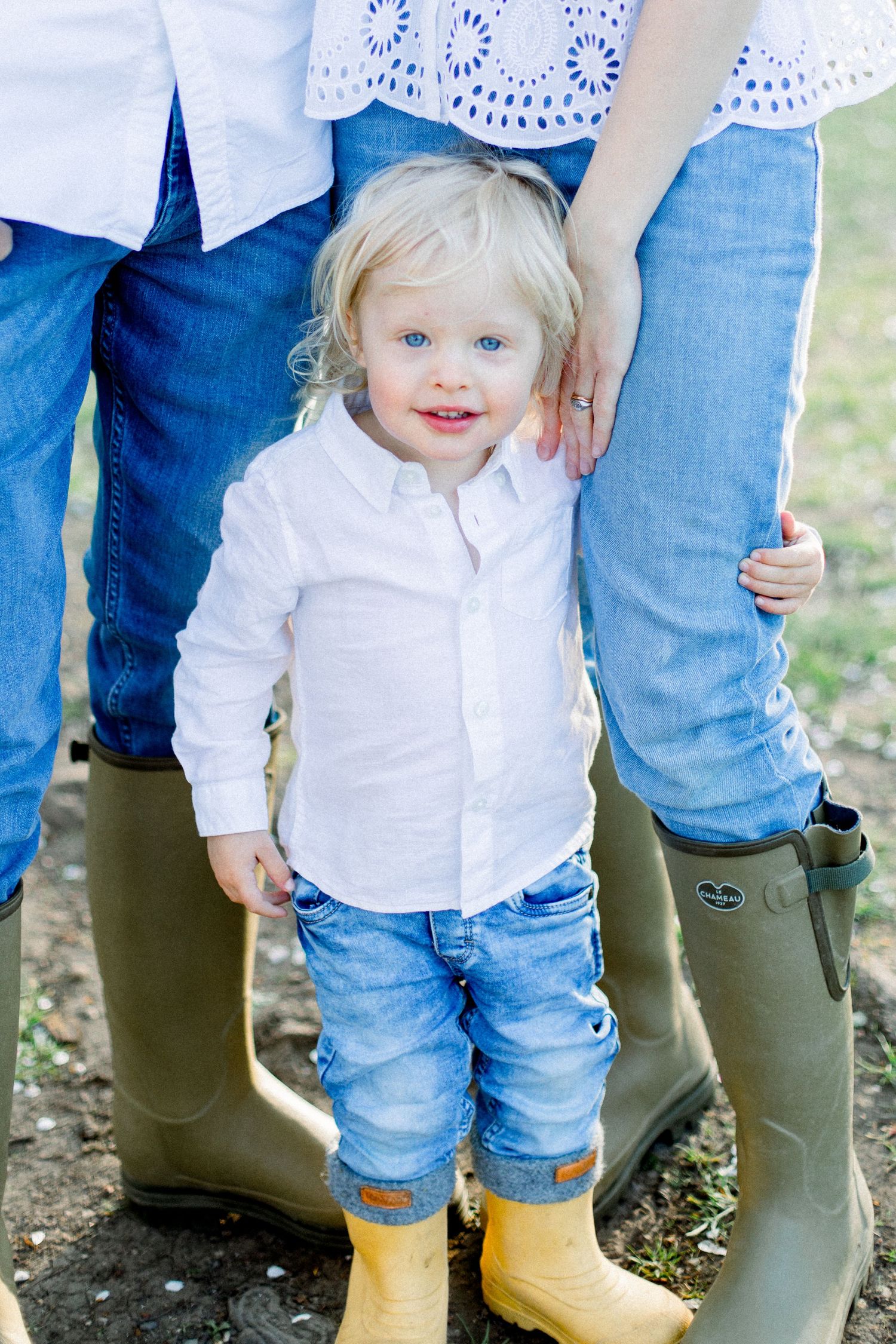 A small child in a white shirt and jeans stands between two pairs of rain boots in an outdoor setting.