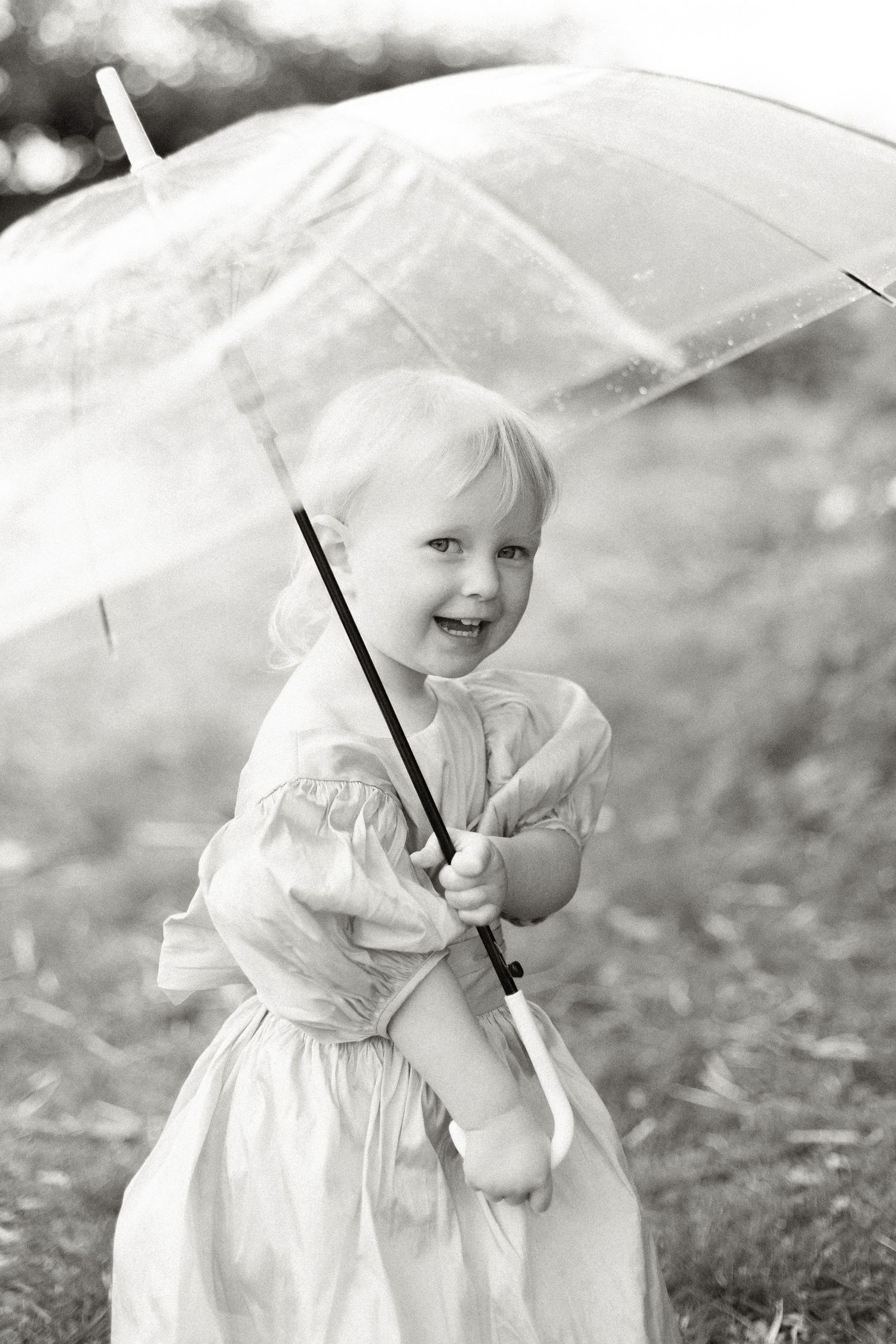 Vintage black and white photo of a child in a white dress holding a white umbrella outdoors.