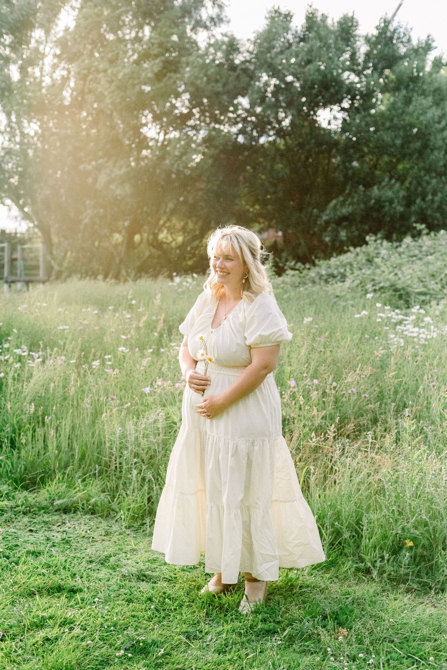 Smiling woman in a flowing cream dress cradles her baby bump while standing in a sunlit meadow of tall grass and wildflowers.